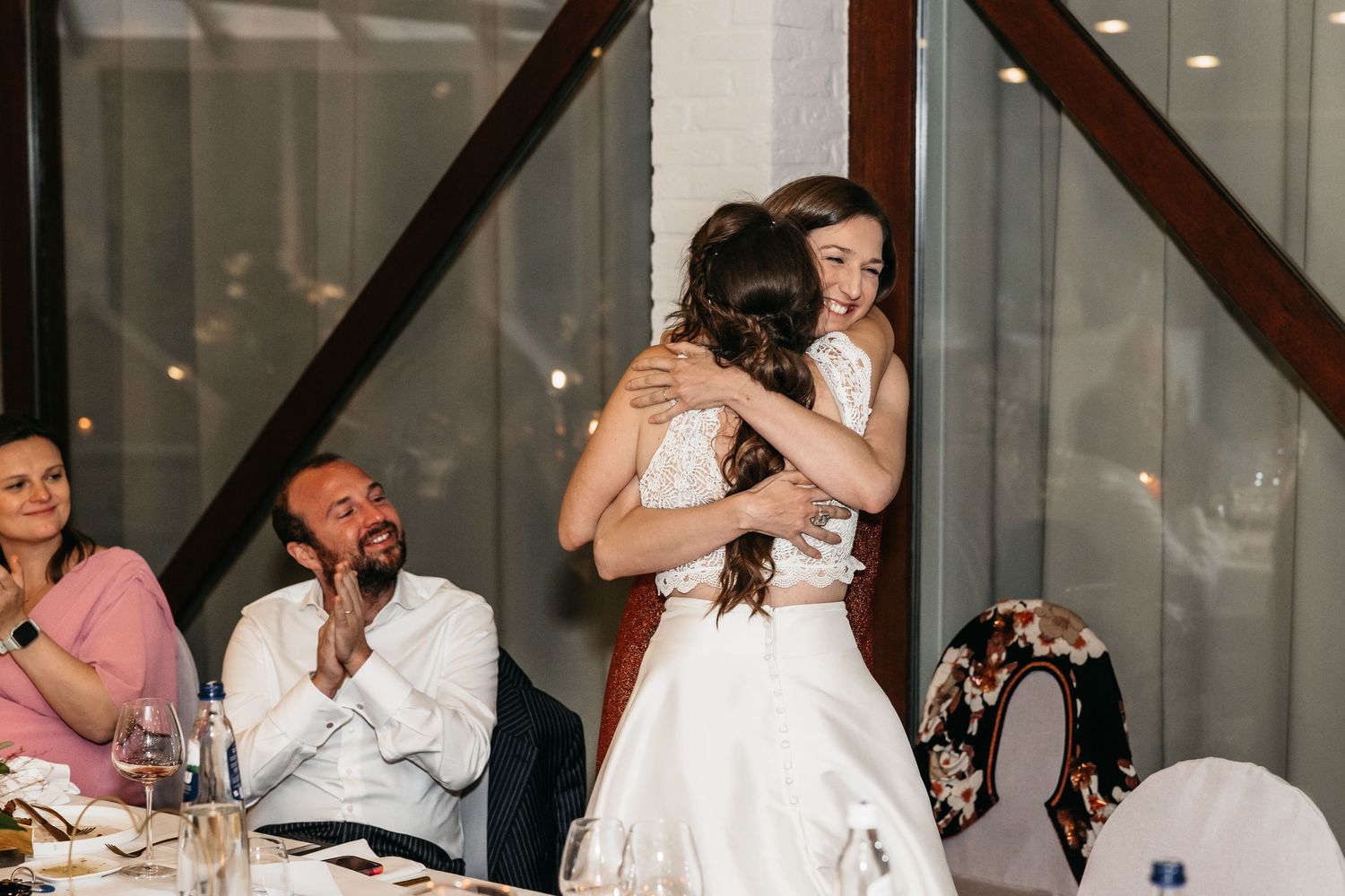 Bride and bridesmaid sharing an emotional hug during wedding reception speech.
