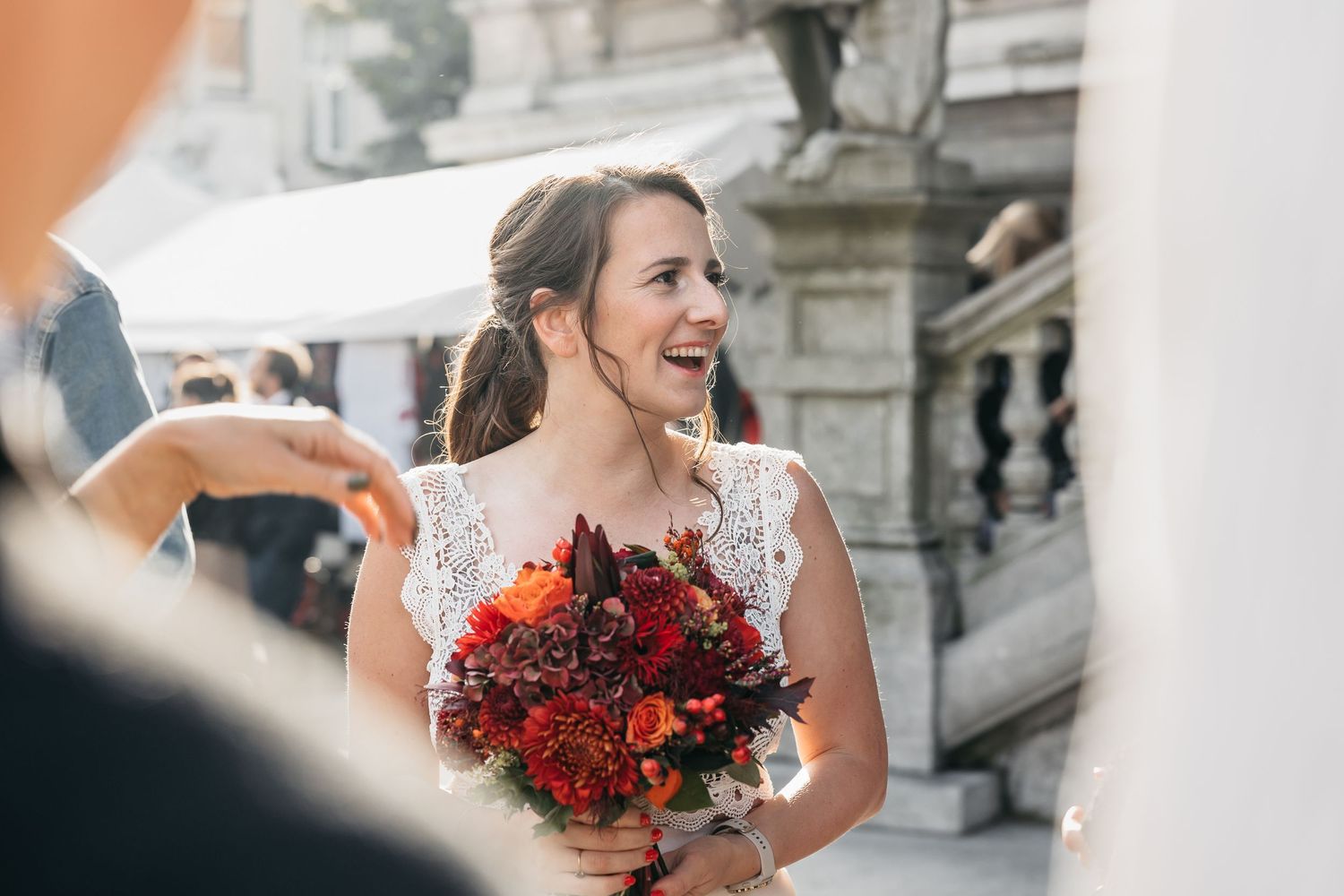 Bride in lace wedding dress laughing joyfully while holding a vibrant red and orange bouquet outdoors.