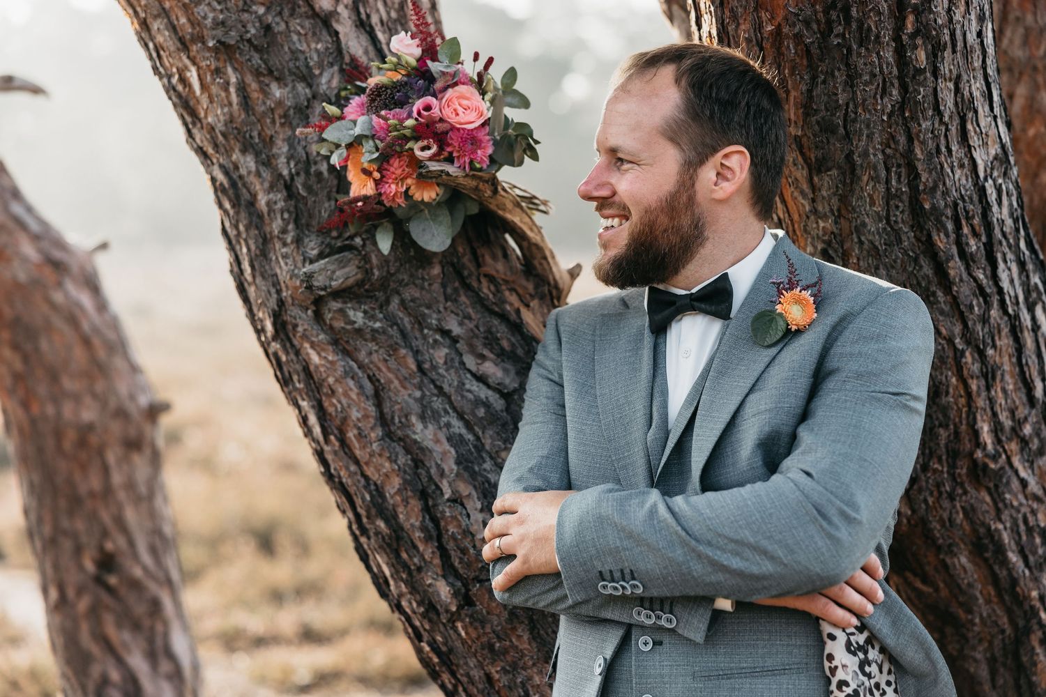 Bearded groom in grey suit and bow tie smiles while leaning against a rustic pine tree with bouquet.