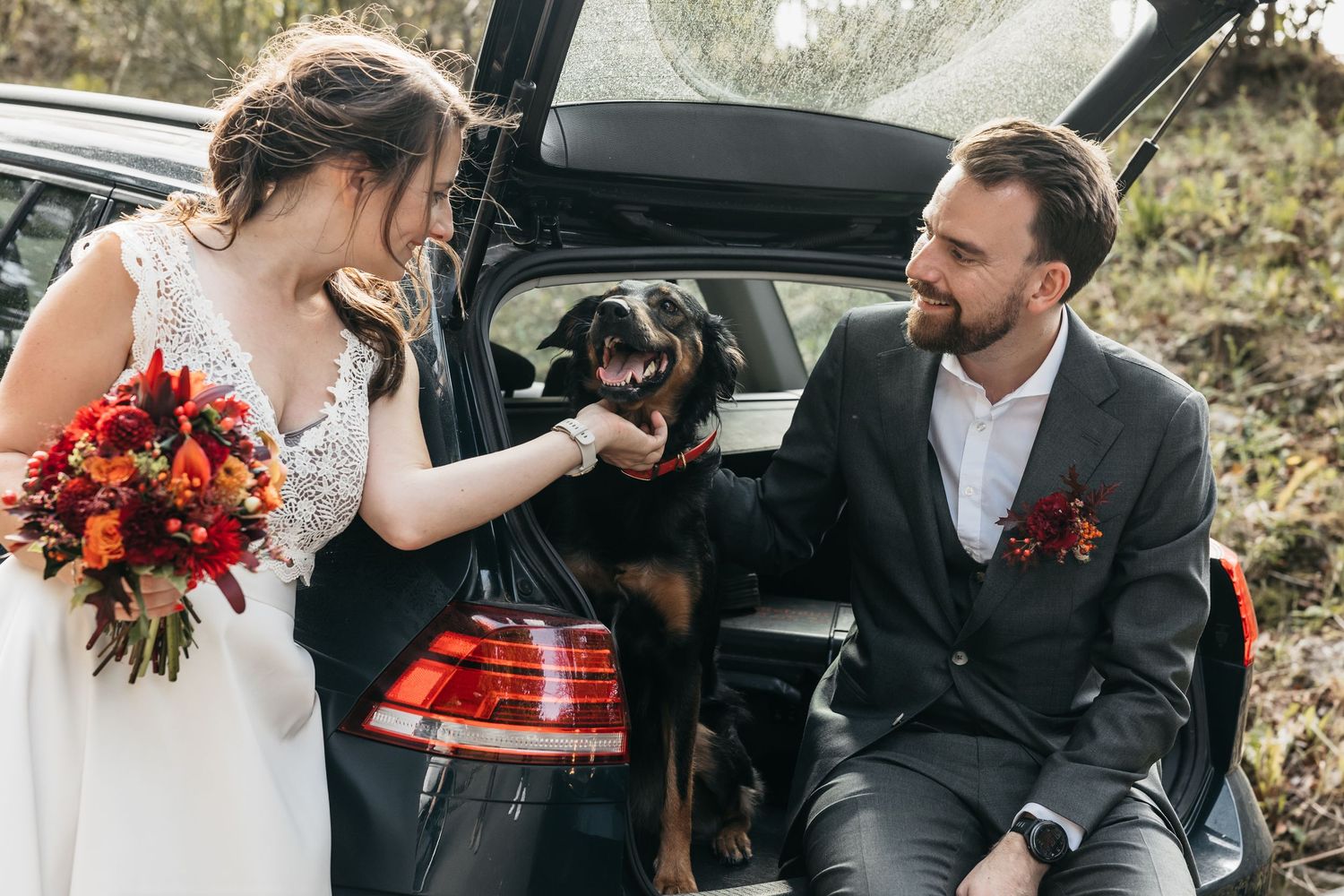 Bride and groom sitting in car trunk with black dog at autumn wedding.