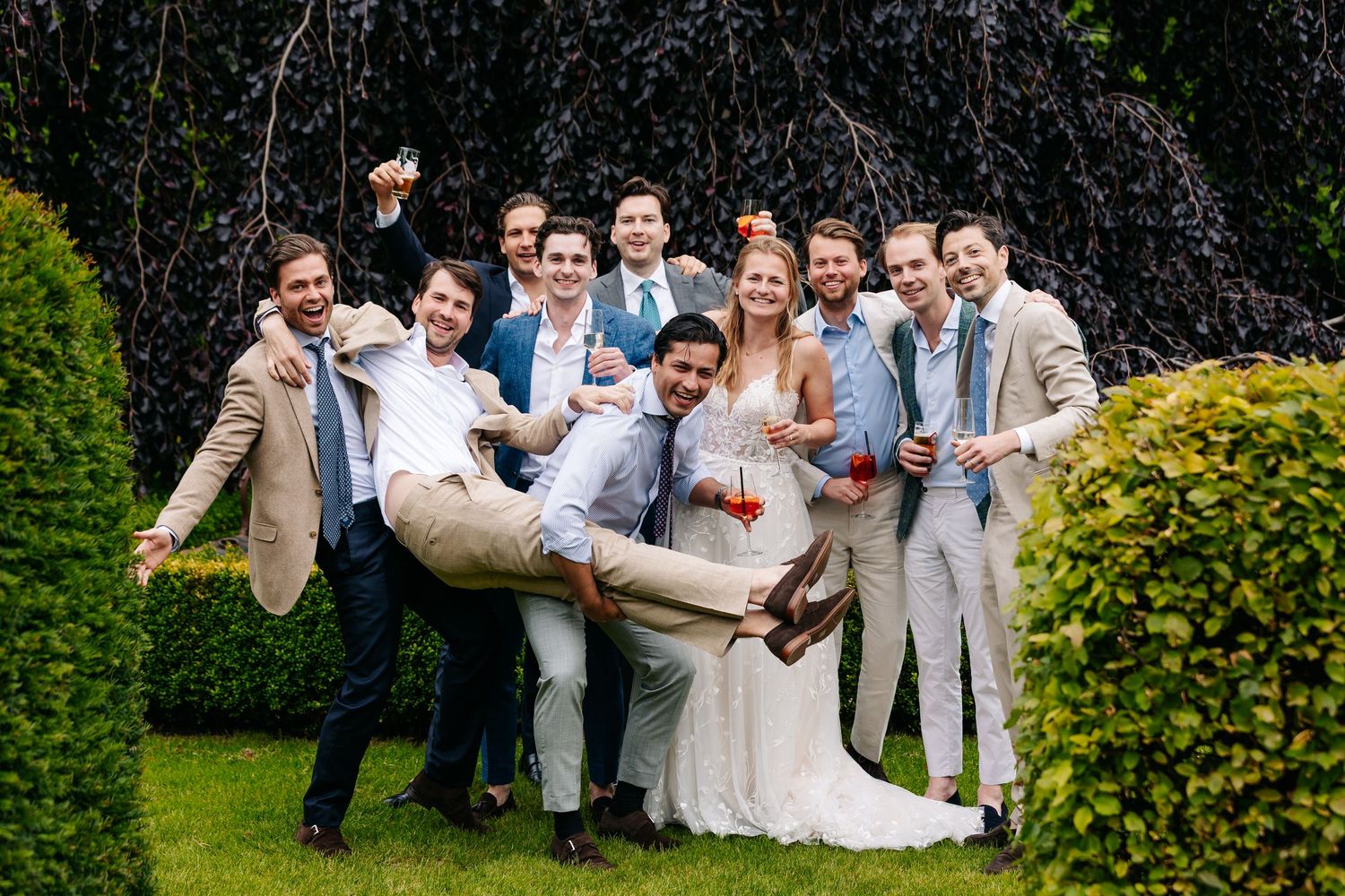 Groom and groomsmen in tan suits lift bride playfully in lush garden setting.