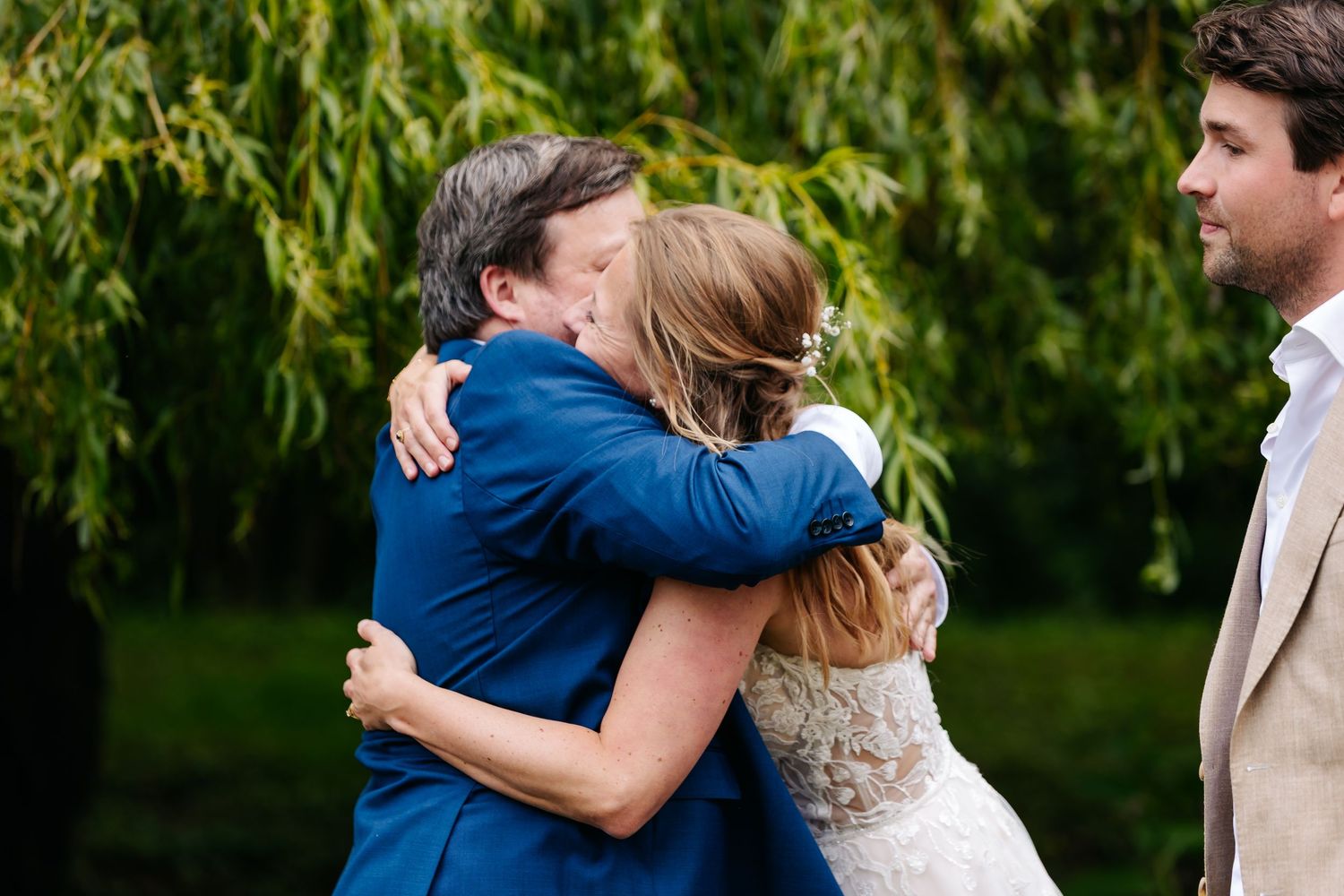 Bride shares an emotional hug with a man in a blue suit under a willow tree.