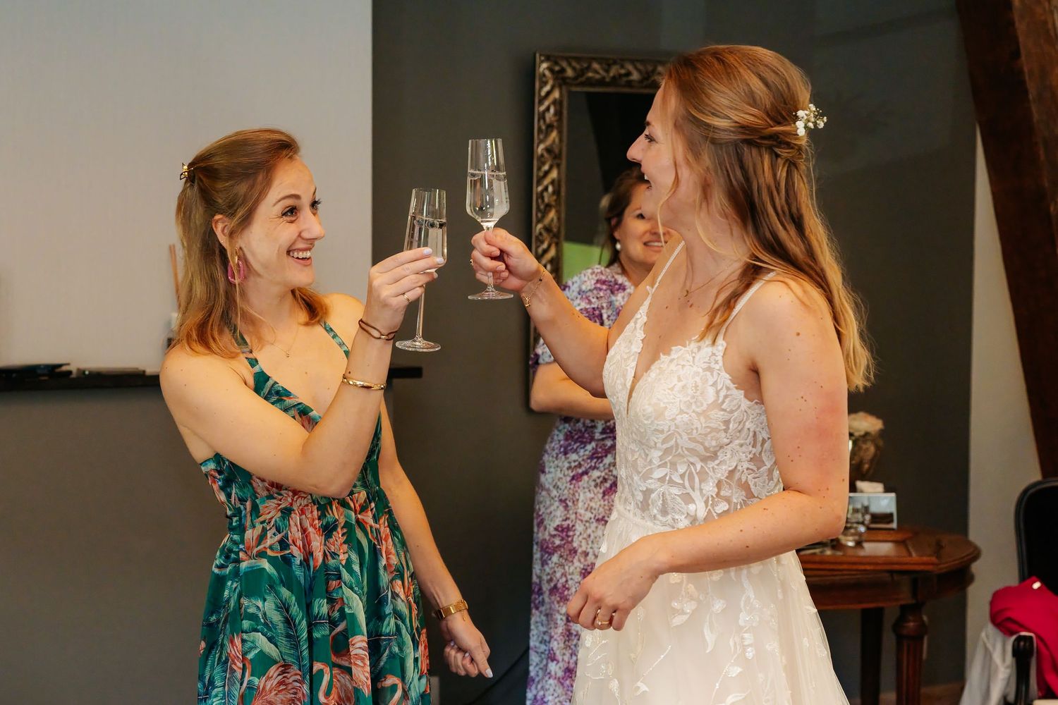 Bride in white lace gown toasting champagne glasses with bridesmaid in floral dress.
