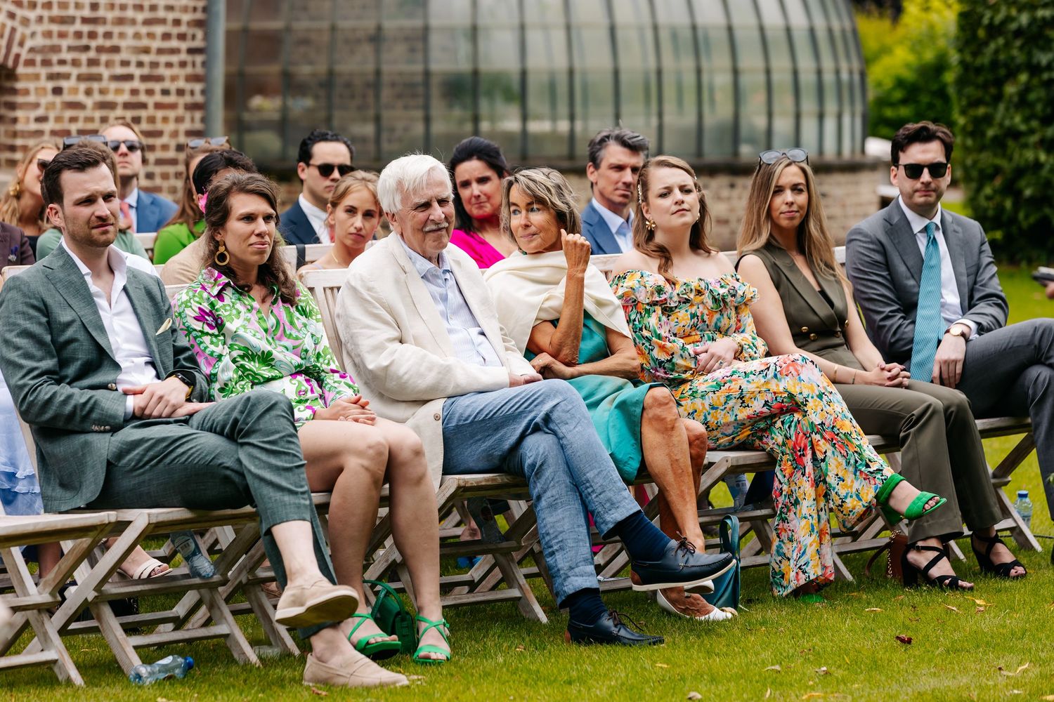 Wedding guests seated outdoors on wooden benches enjoying a sunny garden ceremony in colorful summer attire.