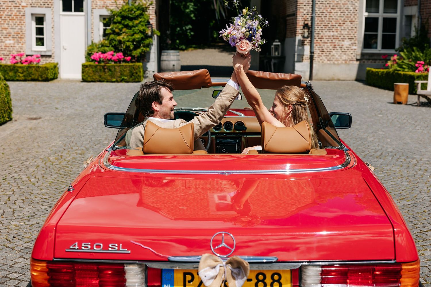 Couple in a red Mercedes 460SL convertible raising a floral bouquet together.