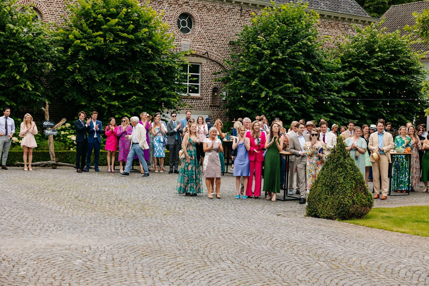 Large wedding party gathering on cobblestone courtyard outside historic brick venue.