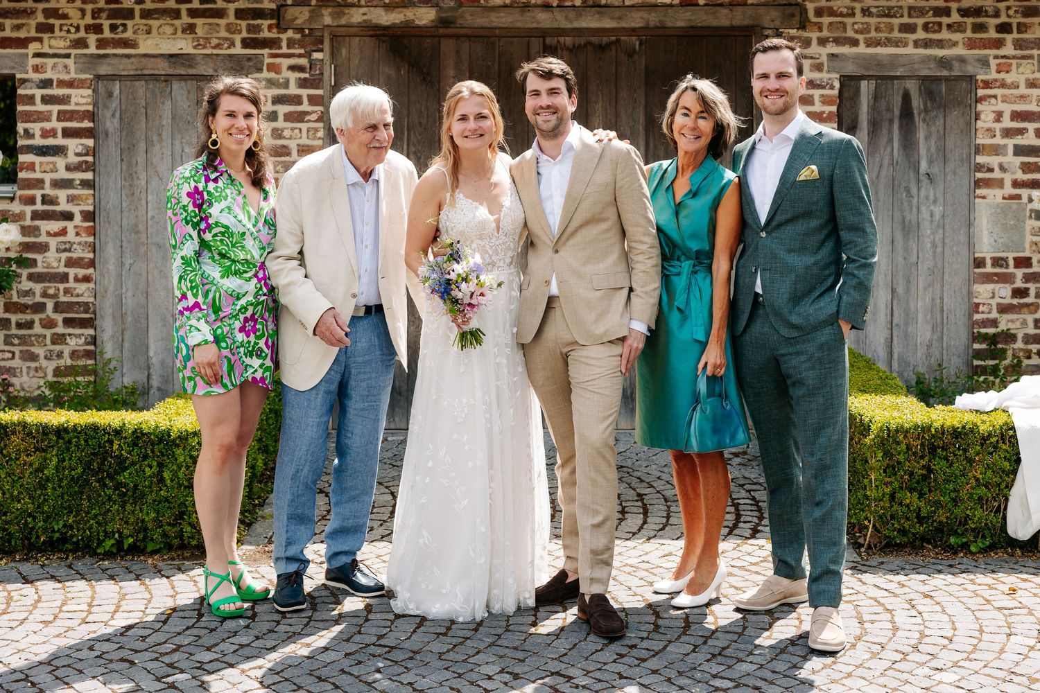 Wedding party of six posing together outdoors, bride in white gown holding bouquet, groom in tan suit, guests in colorful attire.