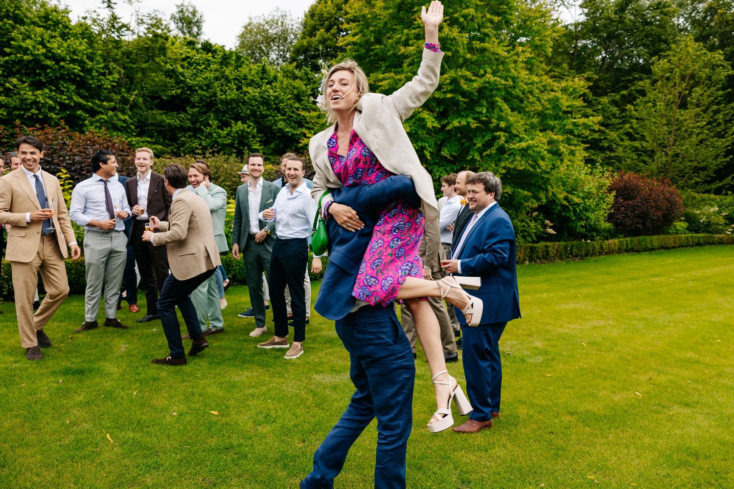 Wedding guest lifted in air by man in navy suit while guests watch on lawn.