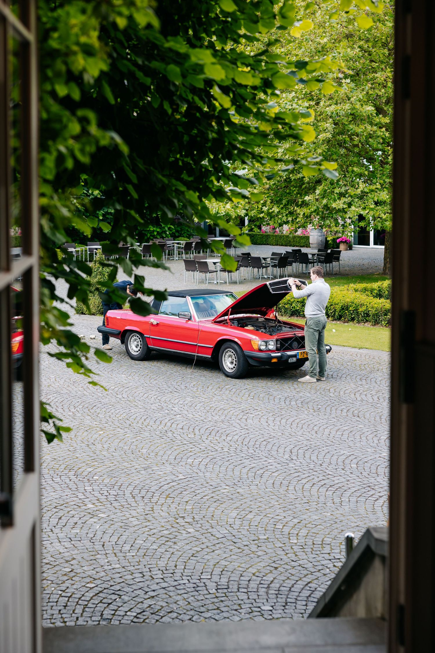Man photographing a classic red convertible Mercedes with open hood on cobblestone driveway.