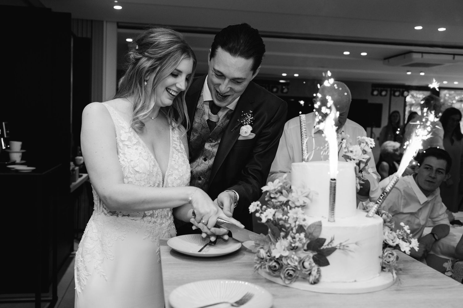 Black and white photo of bride and groom cutting wedding cake with sparklers at reception.