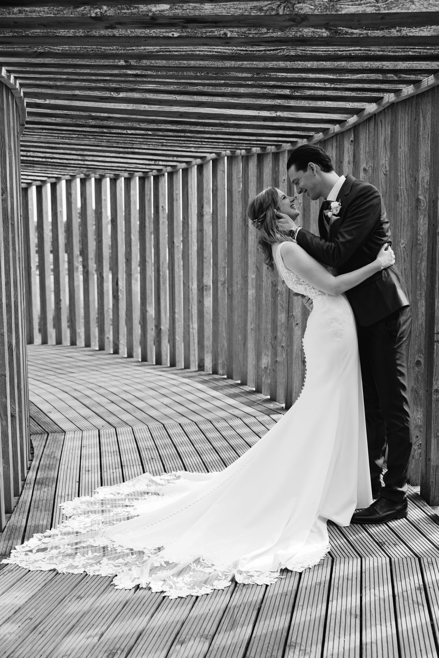 Black and white photo of bride and groom kissing in wooden slatted corridor.