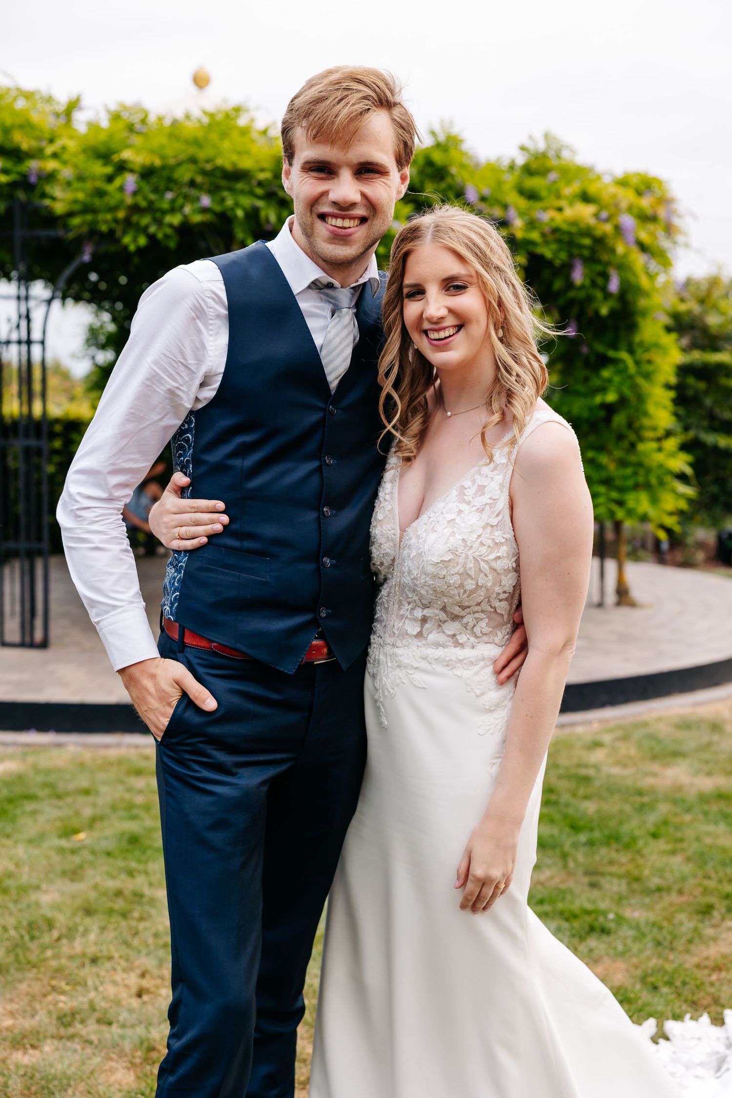 Smiling bride in white lace gown and groom in navy vest pose together in a garden wedding setting.