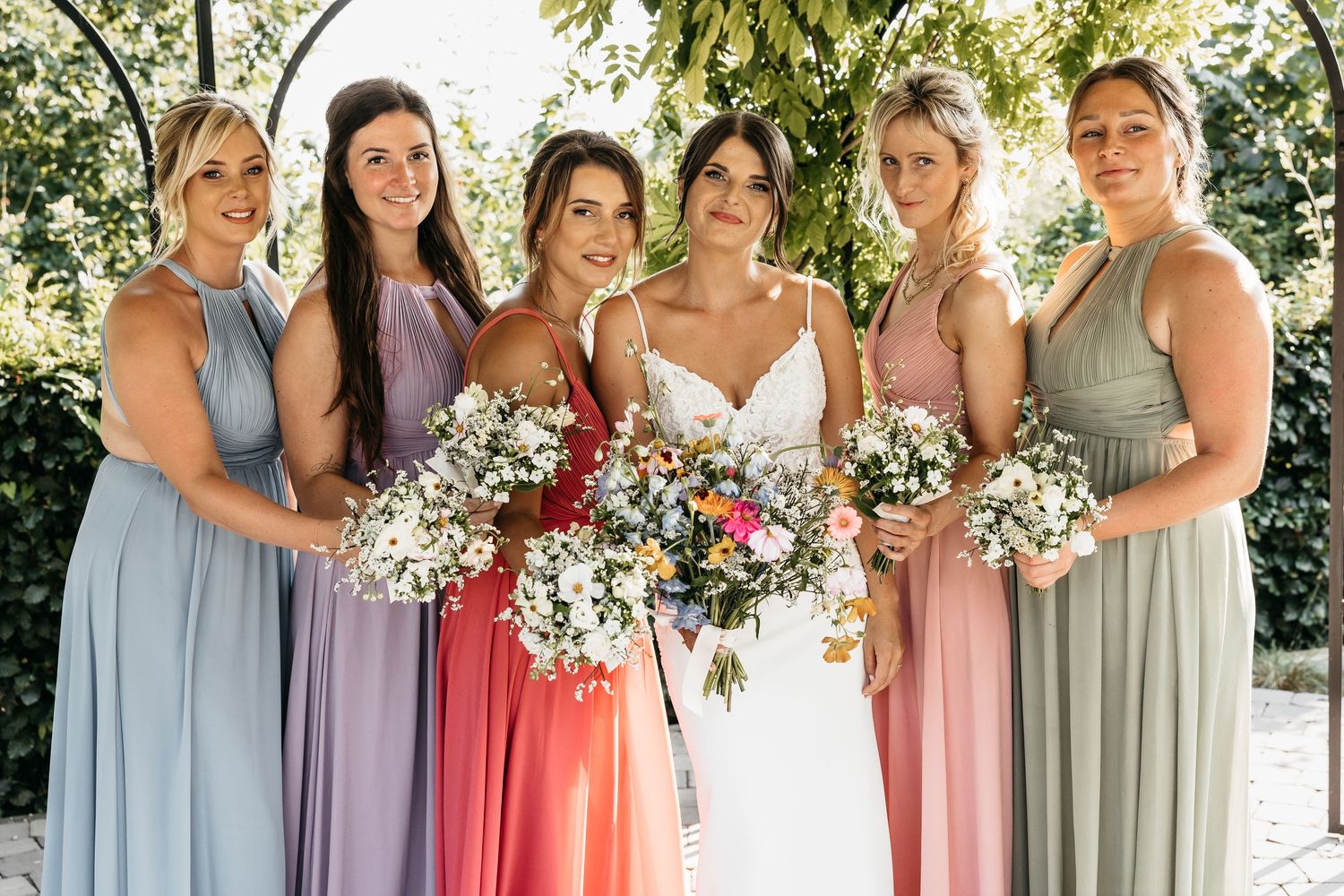 Bride with five bridesmaids in mismatched colorful gowns holding bouquets, posing under a floral arch outdoors.
