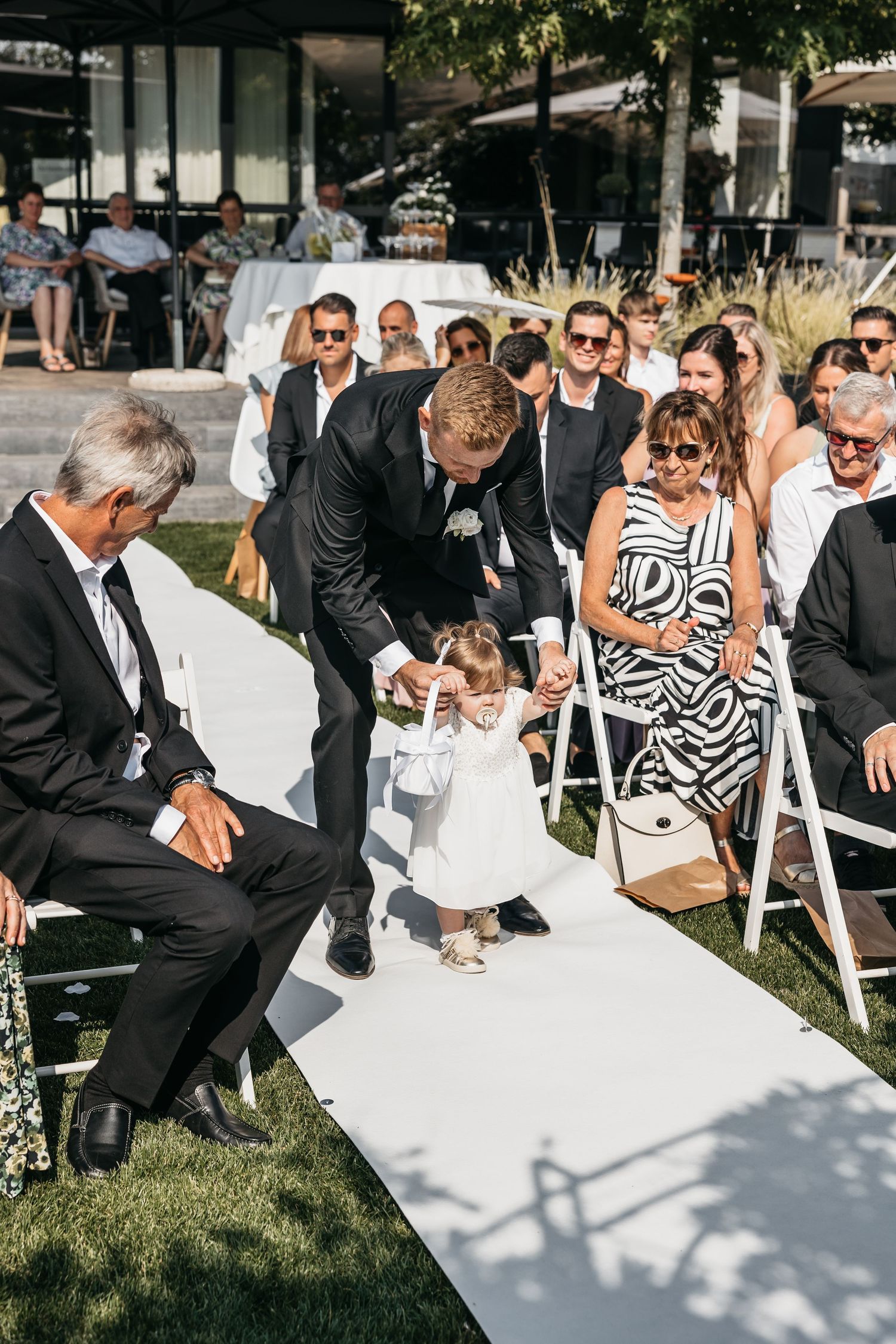 Groomsman helps adorable flower girl in white dress walk down the wedding ceremony aisle.