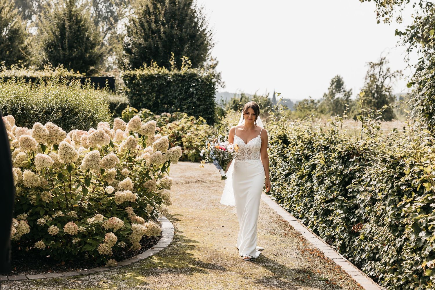 Beautiful bride in white fitted gown carrying bouquet walking along a lush garden path.