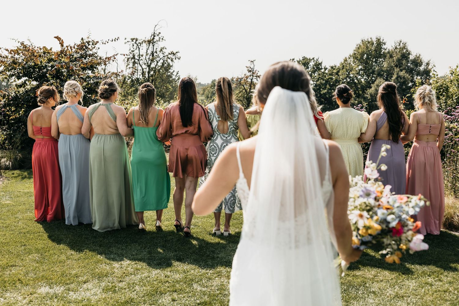 Bride in white gown with veil approaching bridesmaids in colorful dresses standing with backs turned.