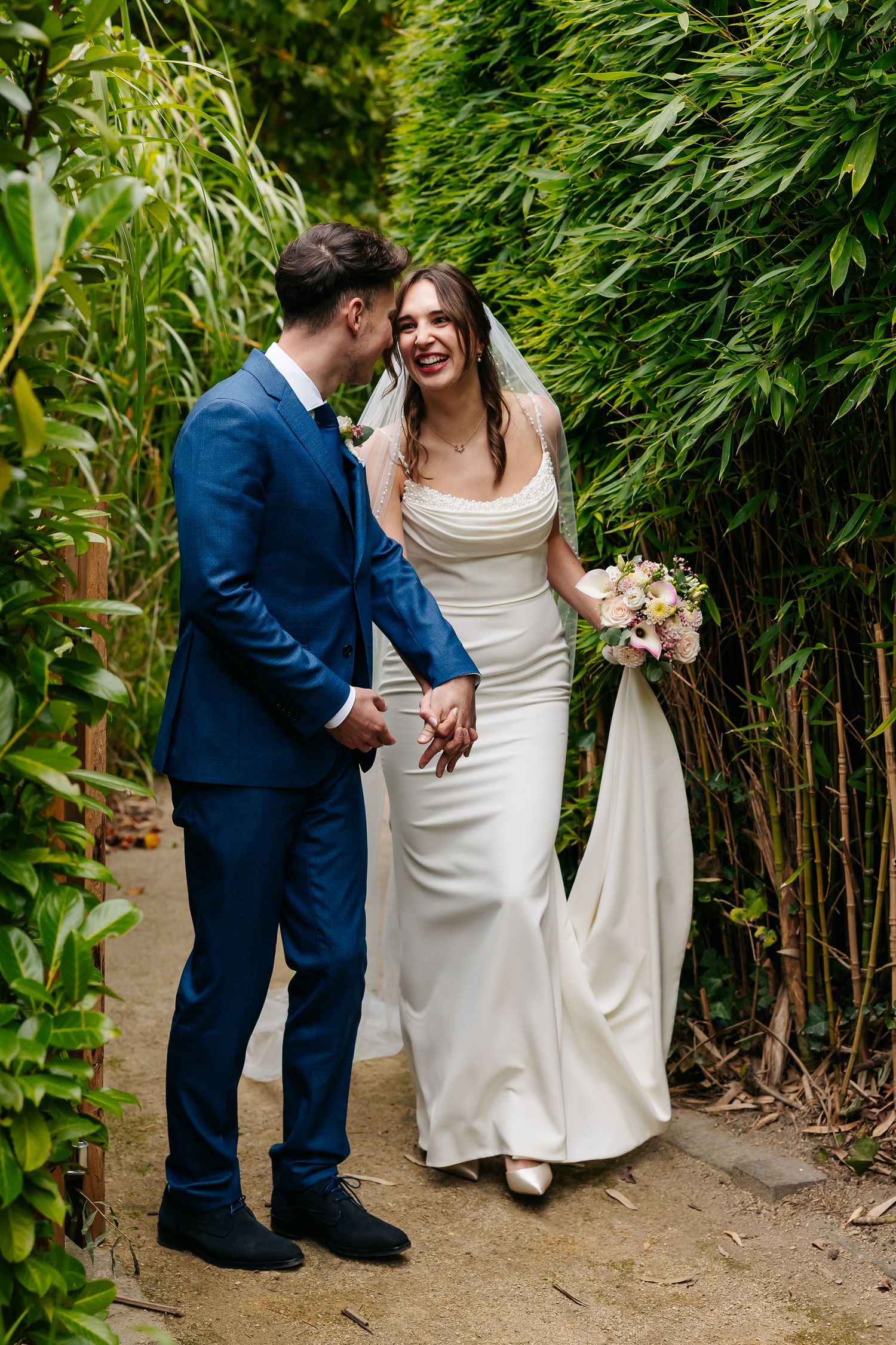 Bride and groom laughing together while walking hand in hand through a lush green bamboo garden.