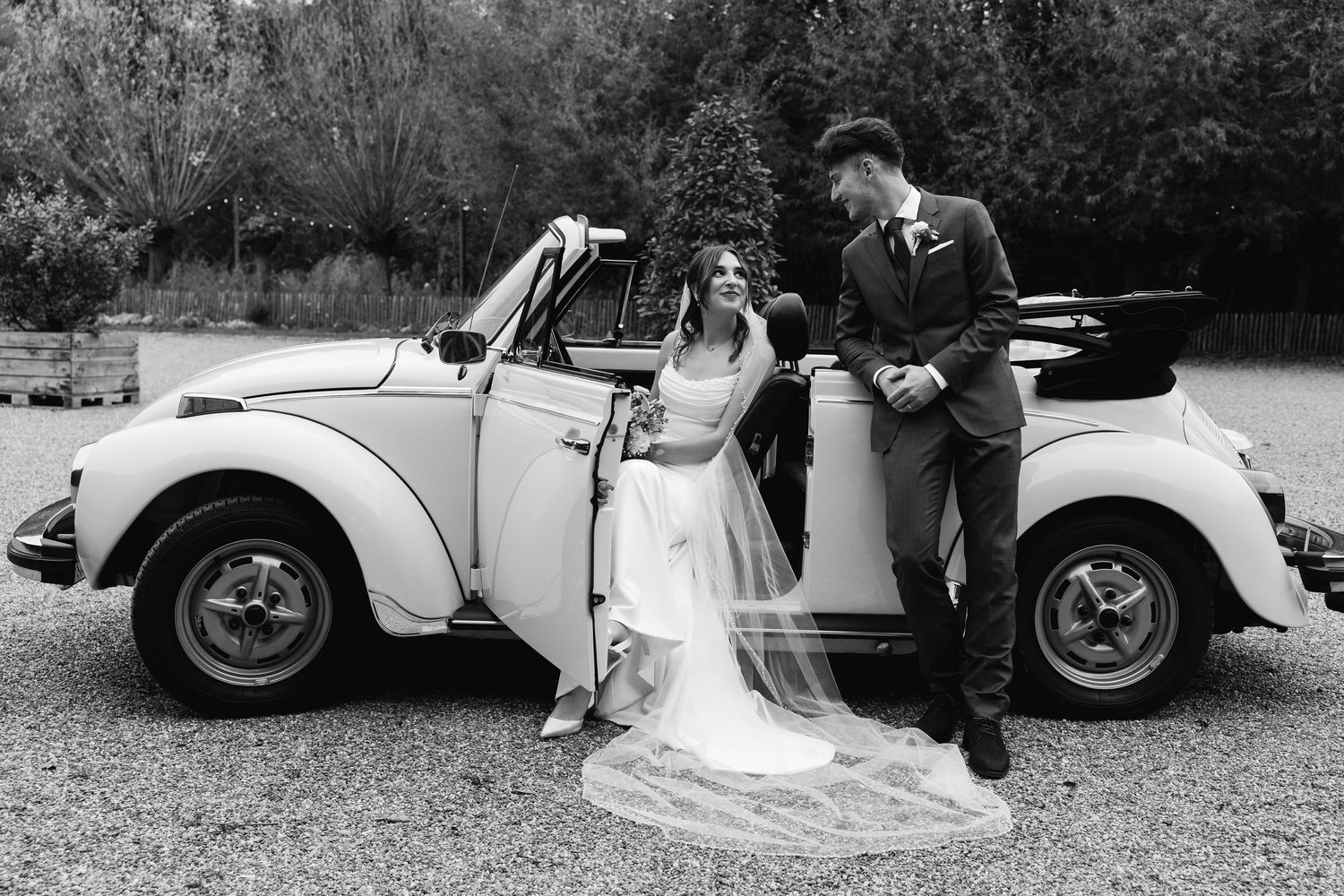 Black and white photo of bride and groom posing beside a classic white VW Beetle convertible wedding car.
