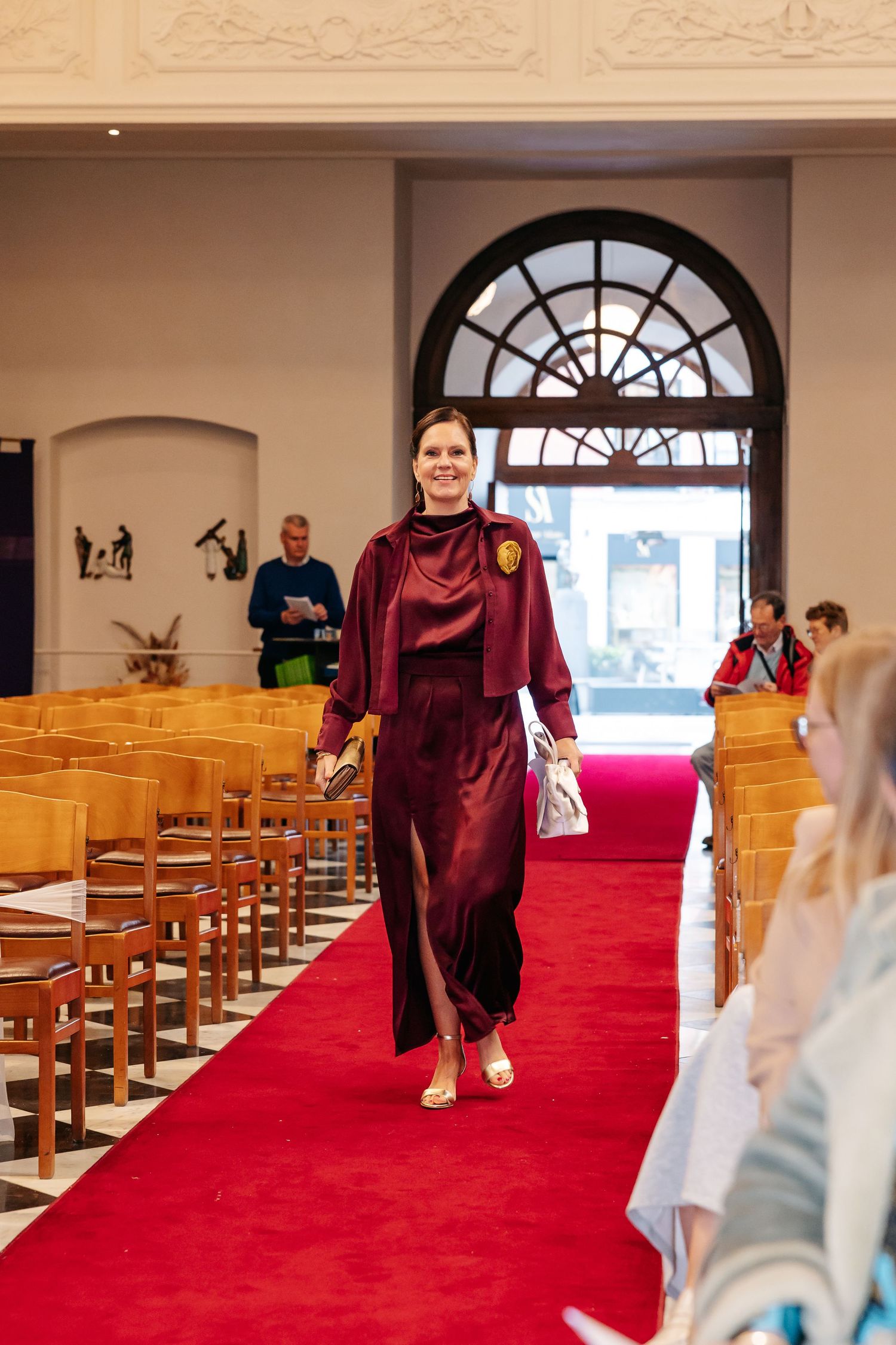 Woman in elegant burgundy velvet gown walking down red carpet aisle inside a beautiful church ceremony.