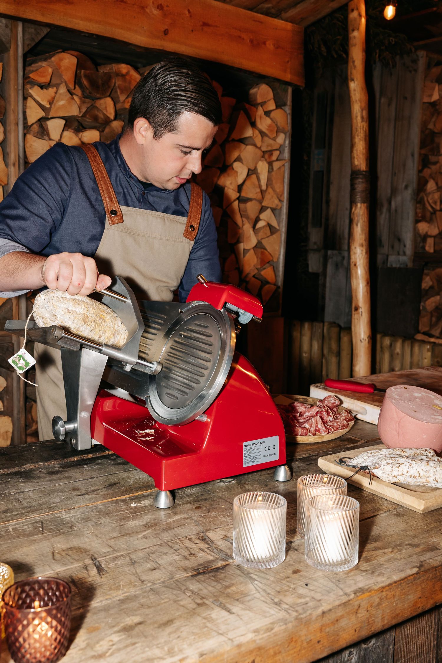 Chef slicing meat on a red electric slicer at a rustic wooden counter with charcuterie.