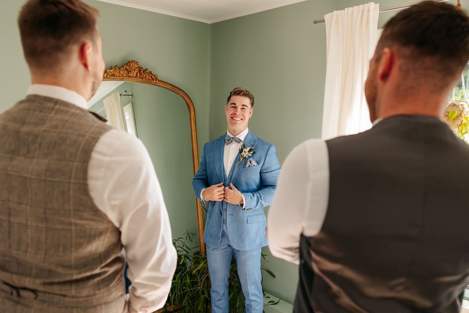 Groom in light blue suit adjusting boutonniere while groomsmen in grey vests look on in green room.