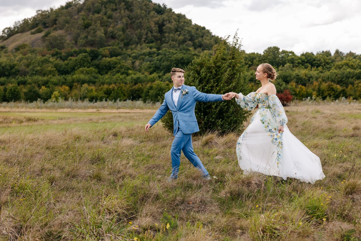 Smiling groom in blue suit leads bride in floral dress by the hand across a grassy field.