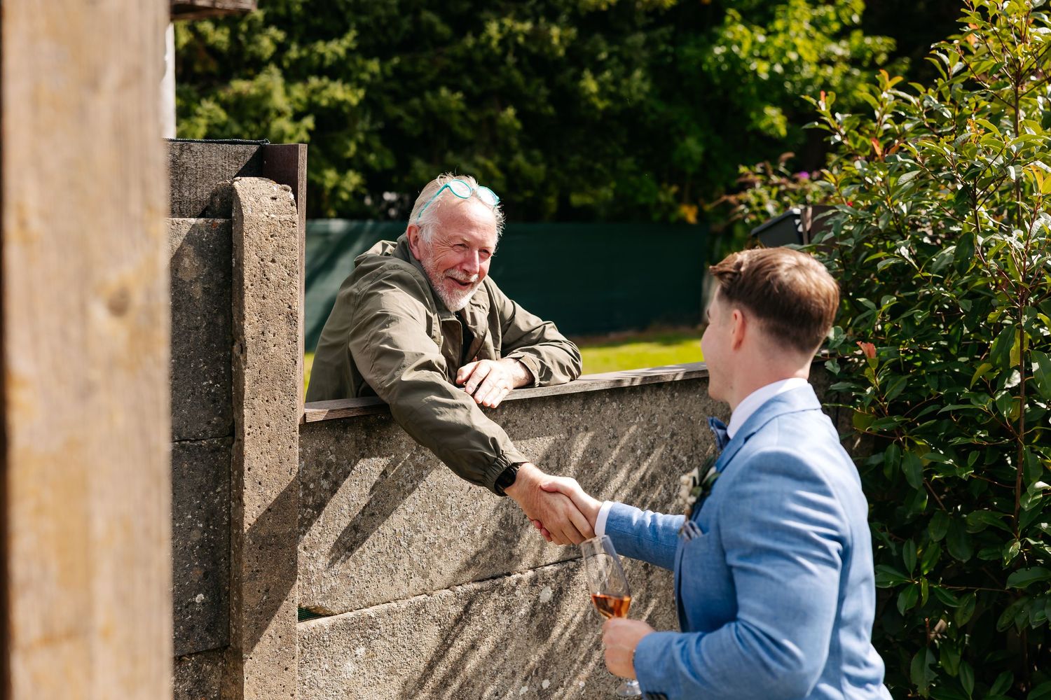 Groom in blue suit shaking hands with elderly man over stone wall outdoors.