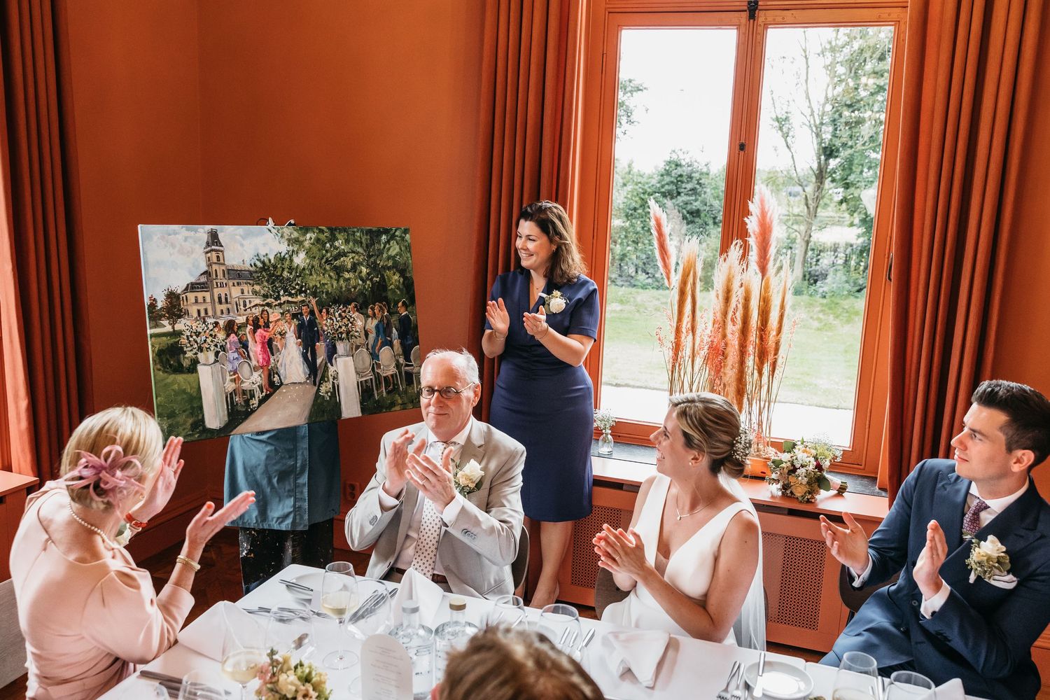 Woman presents large wedding photo canvas to applauding guests at elegant reception dinner.