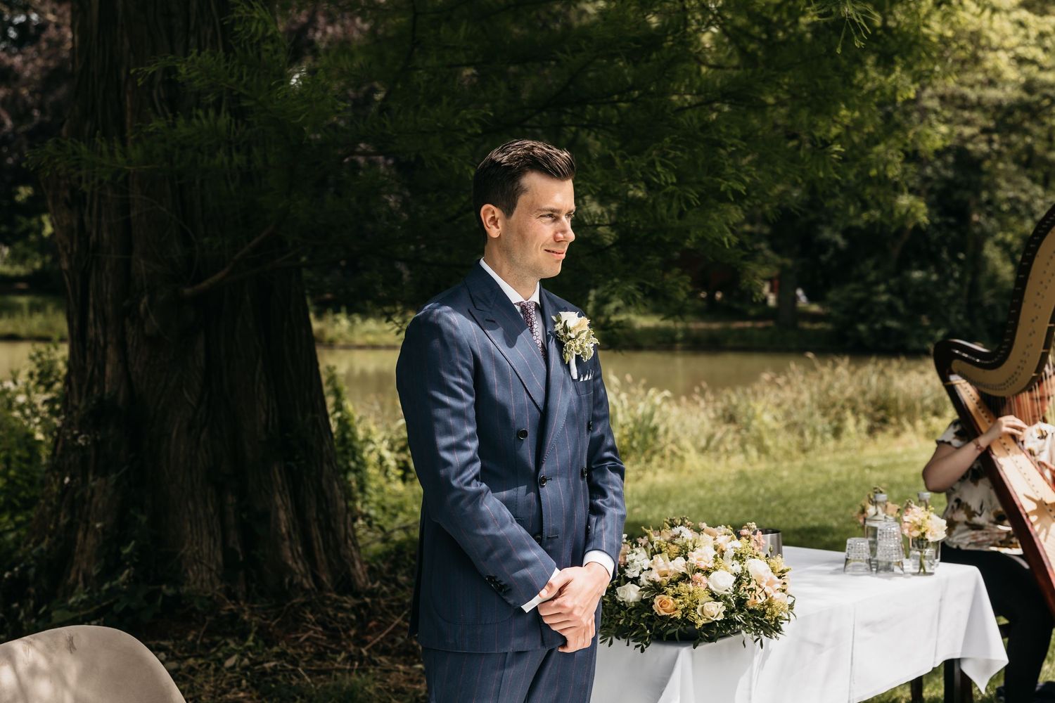 Groom in navy blue suit stands solemnly at outdoor wedding altar surrounded by floral arrangements.