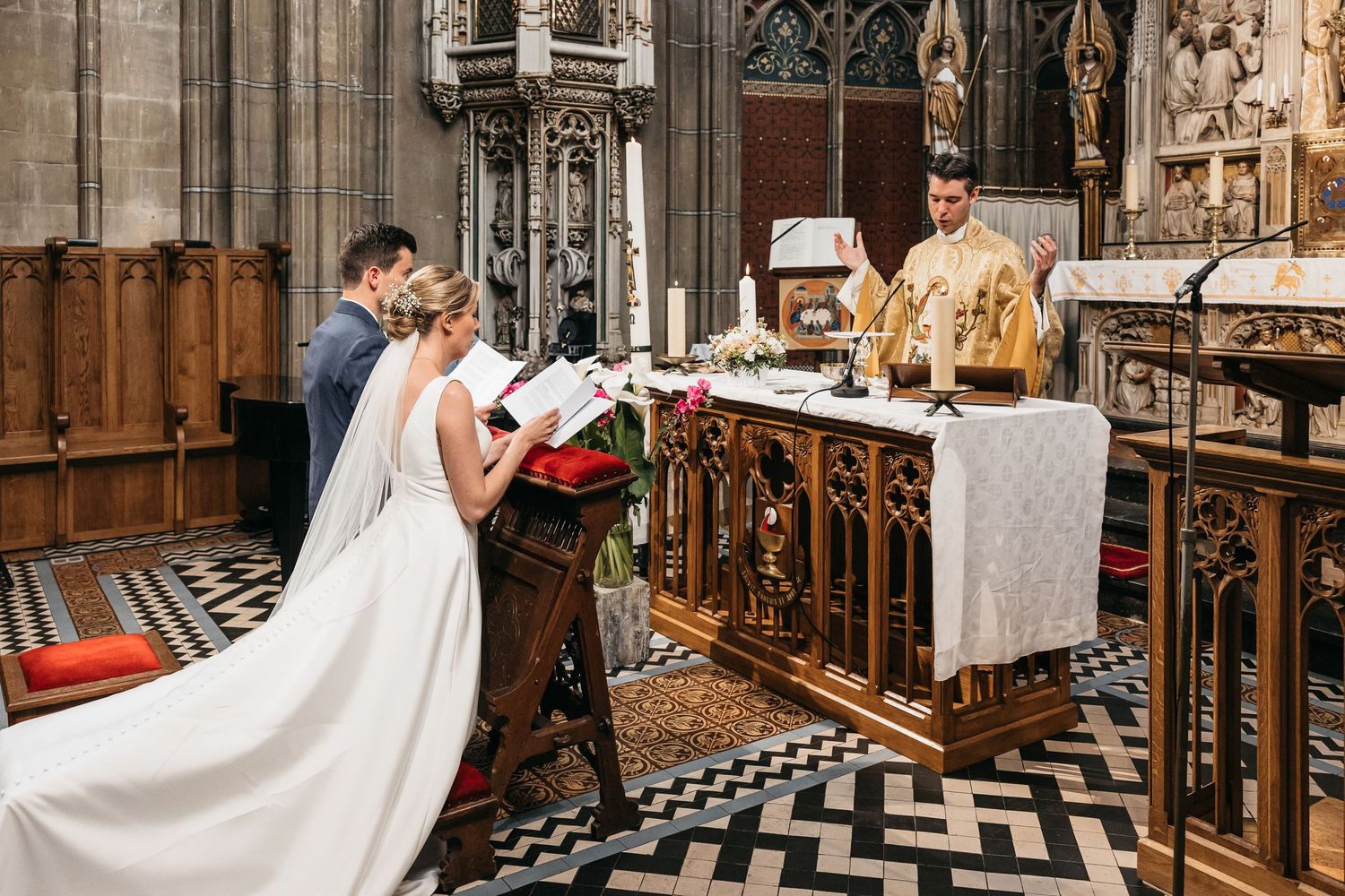 Bride and groom kneel before golden-robed priest at ornate altar during Catholic wedding ceremony.