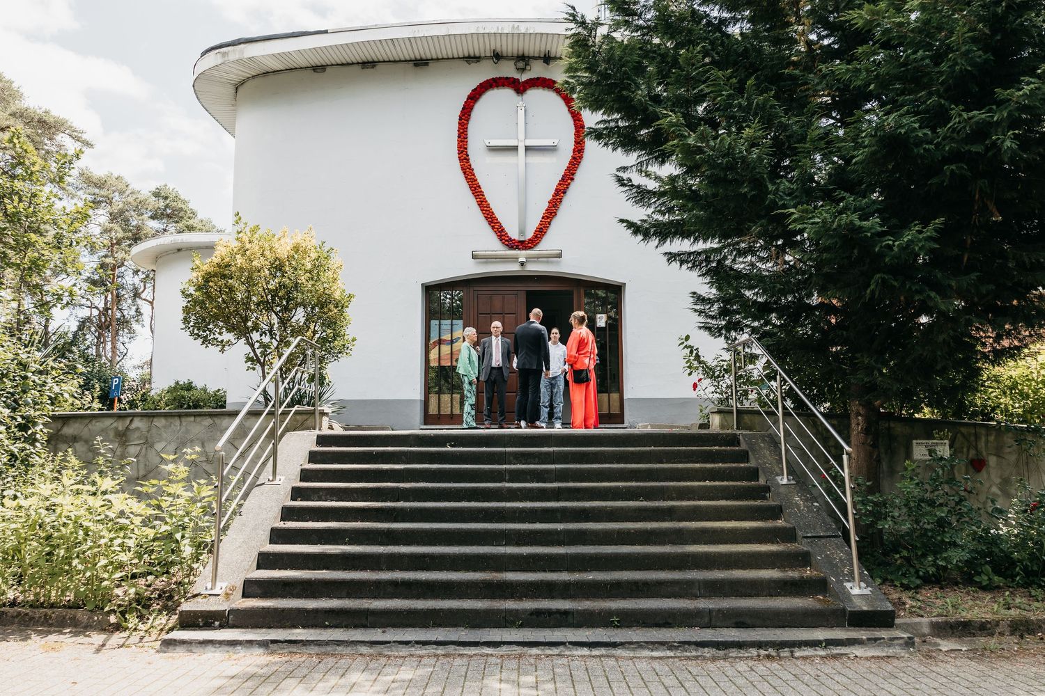 Wedding party posing on steps of a white circular chapel entrance decorated with a large red heart symbol.