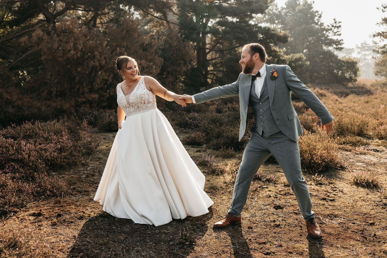 Bride and groom hold hands and dance joyfully in a sunlit autumn heathland during their wedding photo shoot.