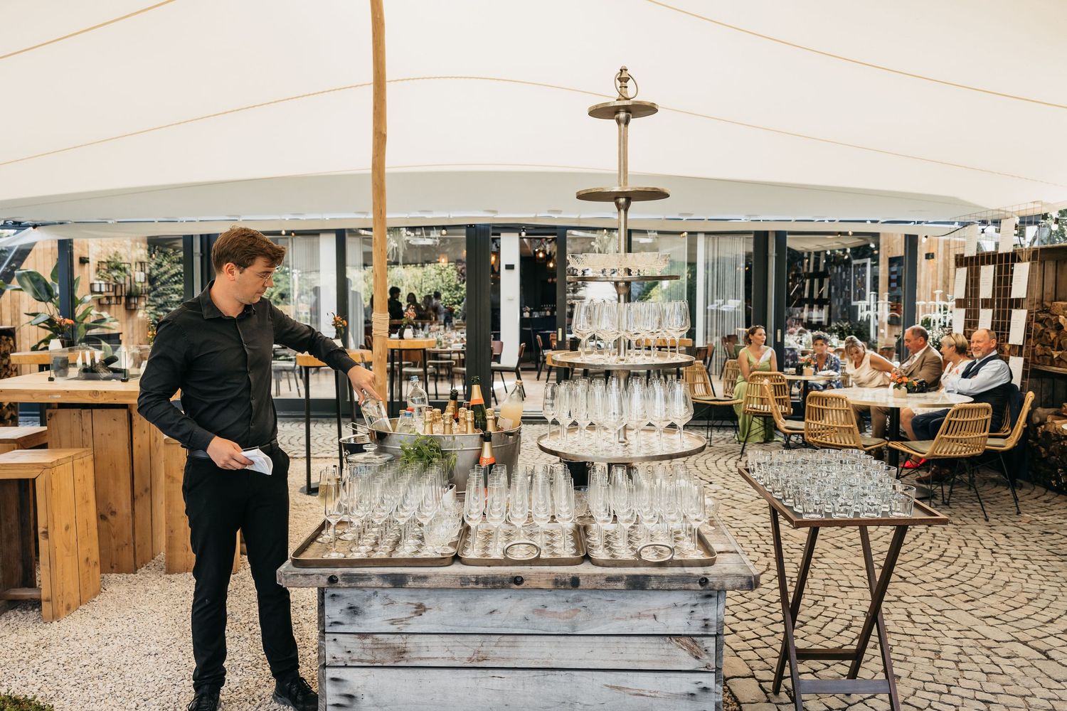 Waiter arranging champagne tower on rustic wooden table under a white marquee at an outdoor wedding reception.