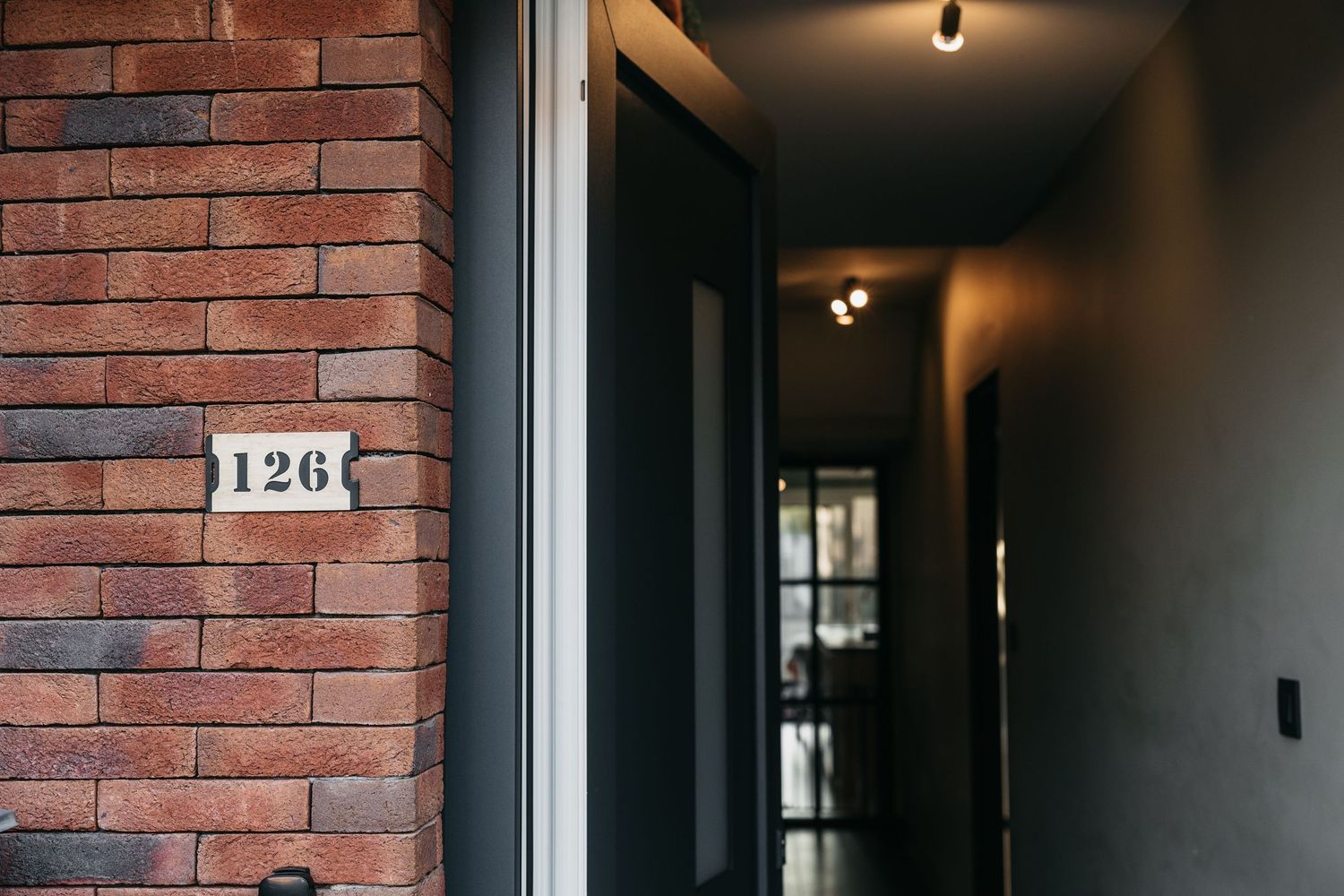 Number 126 address plaque on red brick wall beside a modern dark hallway entrance door.