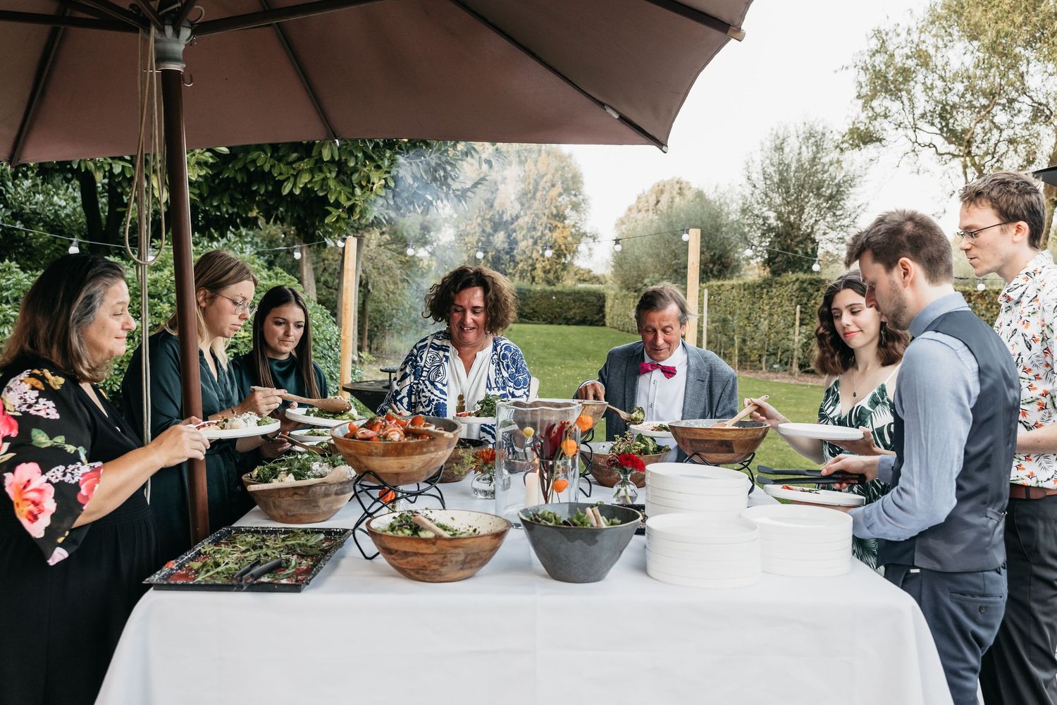 Guests serving themselves from a large outdoor buffet table with salads under a garden umbrella.