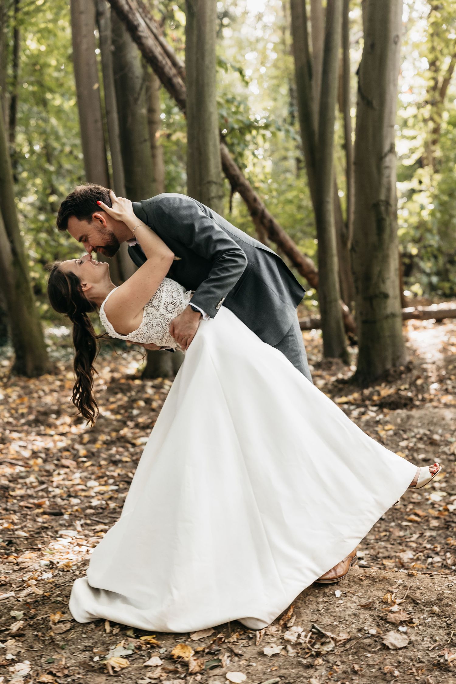 Groom dipping bride in flowing white gown for a passionate kiss among fall forest trees.