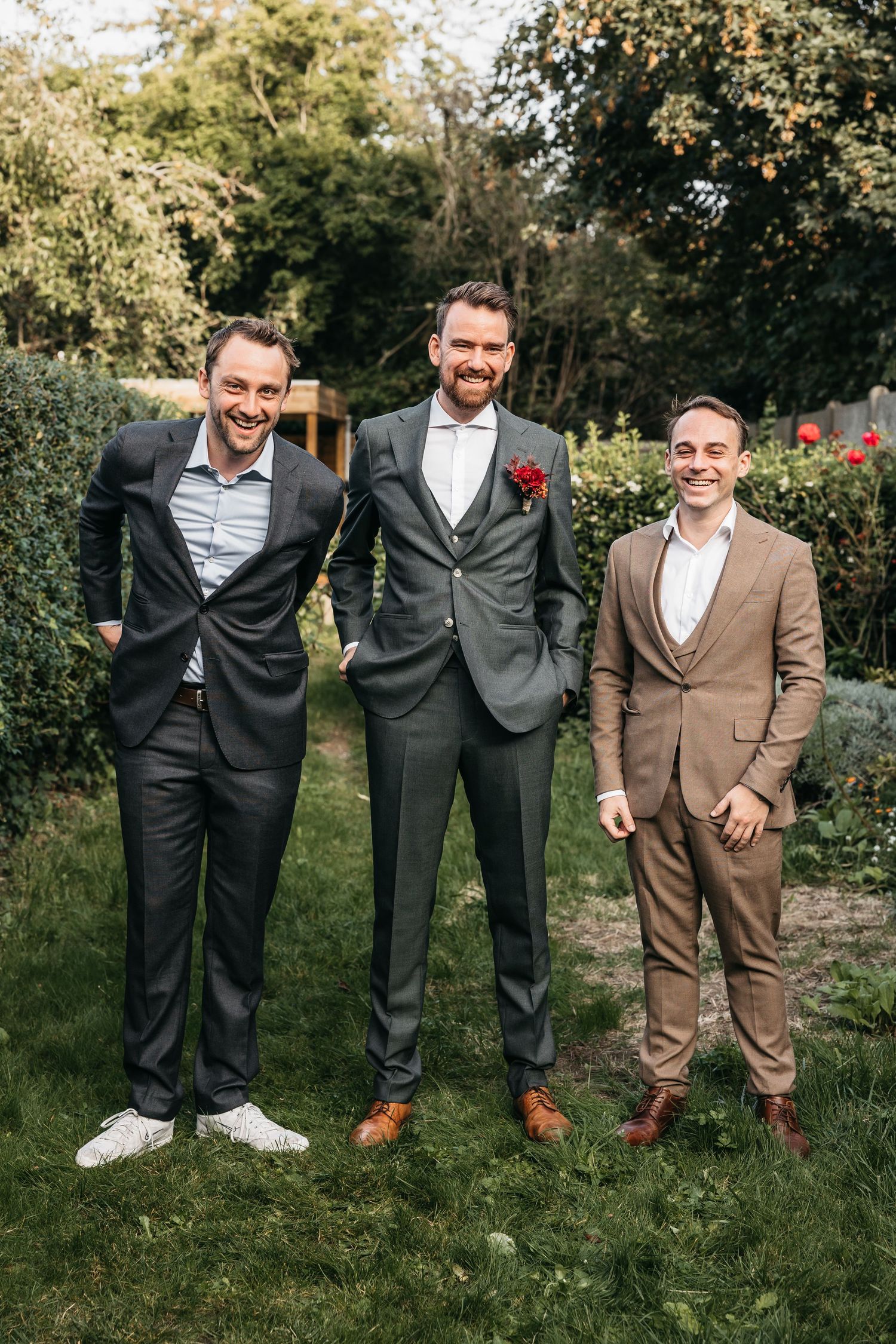 Groom in gray suit with red boutonniere laughing with two groomsmen in dark and tan suits outdoors.