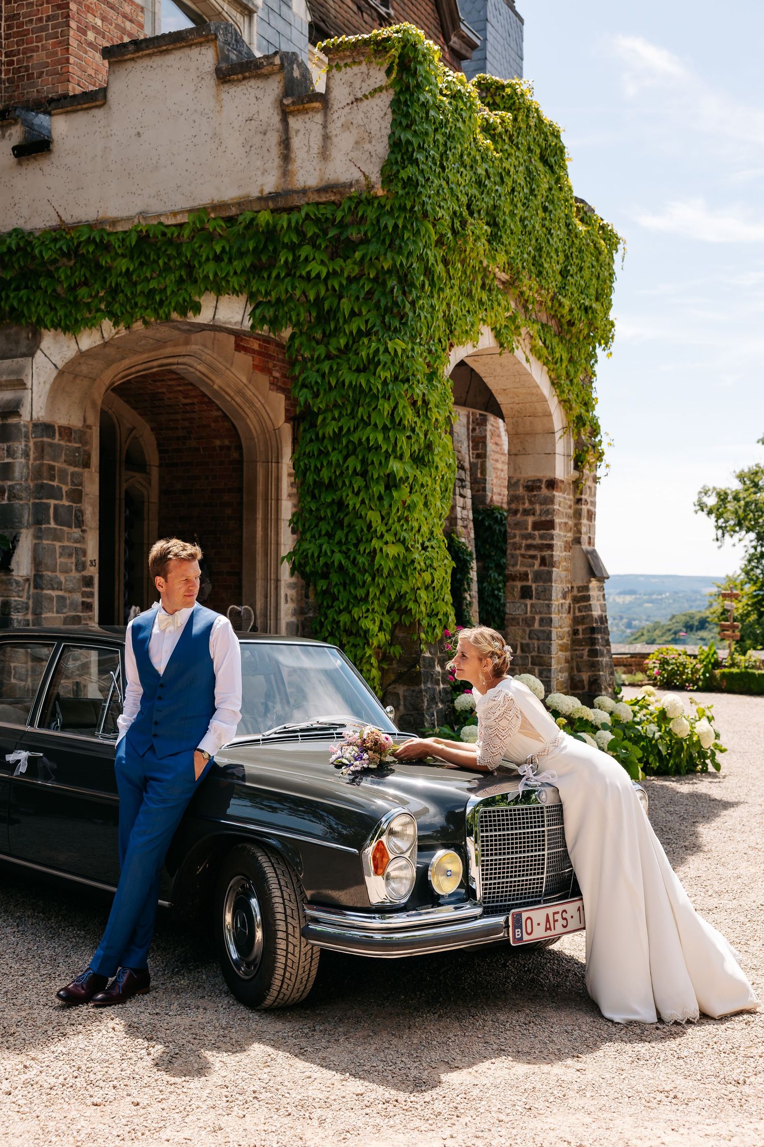 Bride leaning on a vintage black Mercedes while groom stands beside it at an ivy-covered castle.
