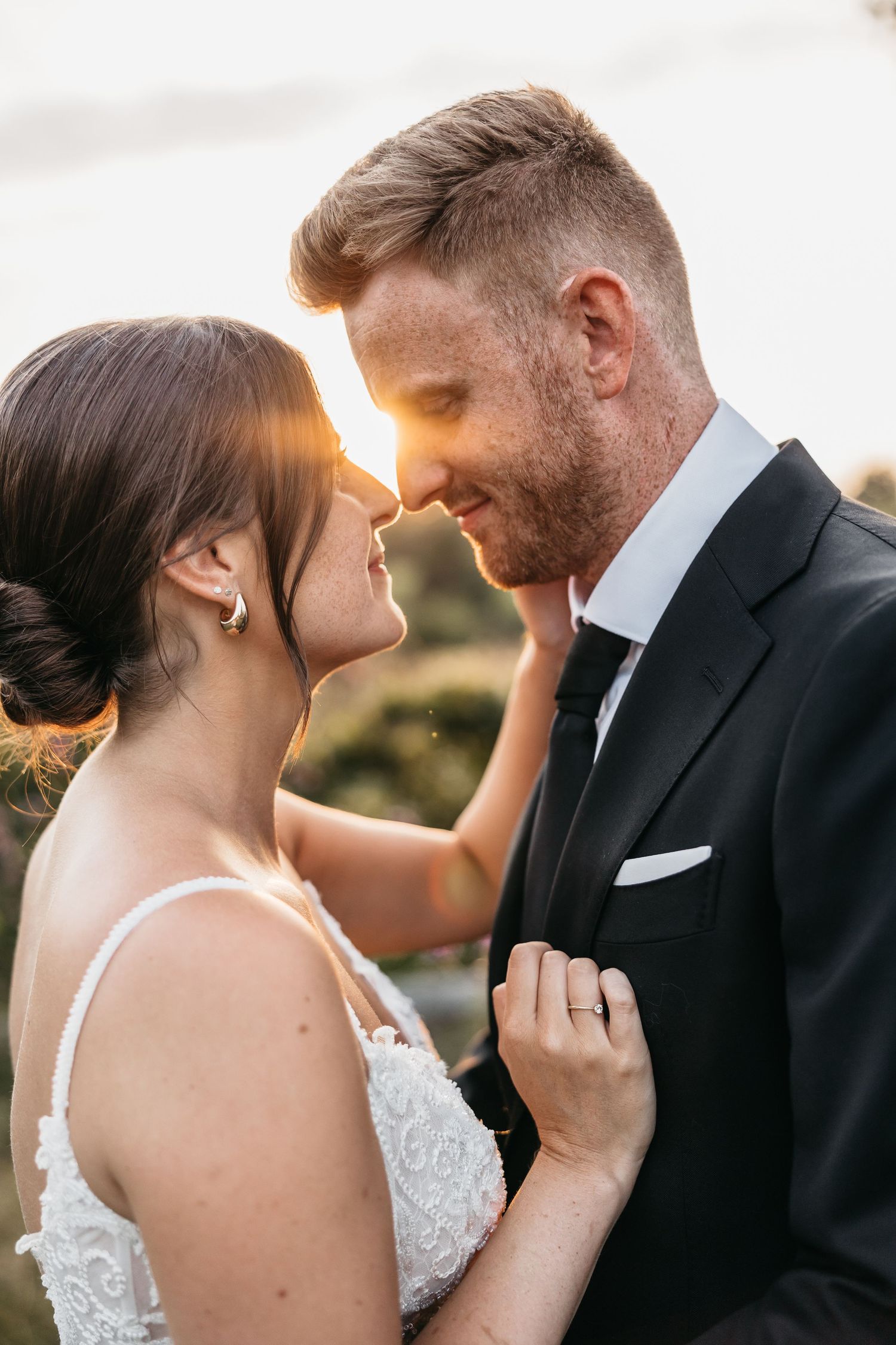 Bride and groom share an intimate nose-to-nose moment bathed in warm golden sunset light outdoors.