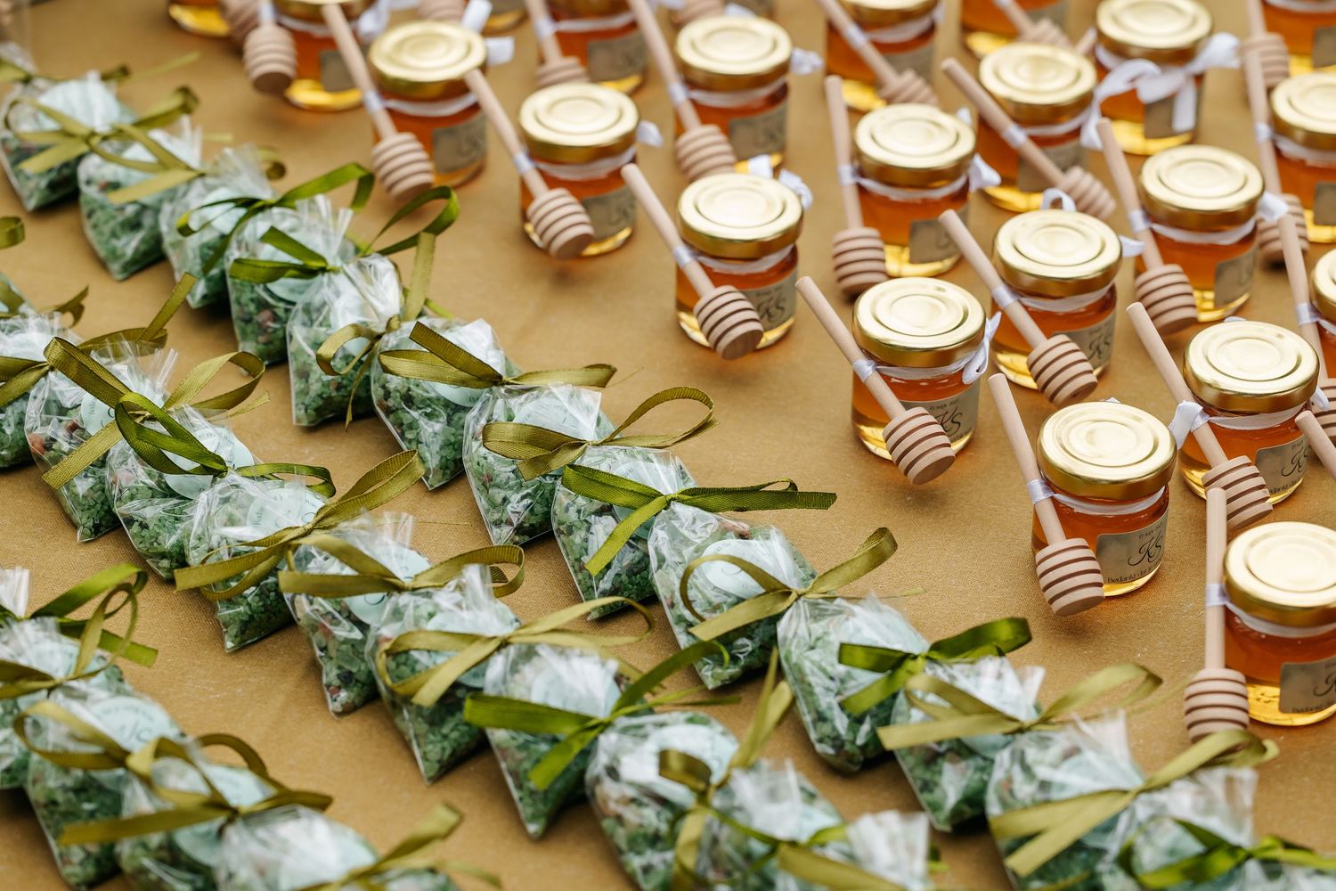 Rows of honey jar wedding favors with wooden dippers and herb bundle gifts on display table.
