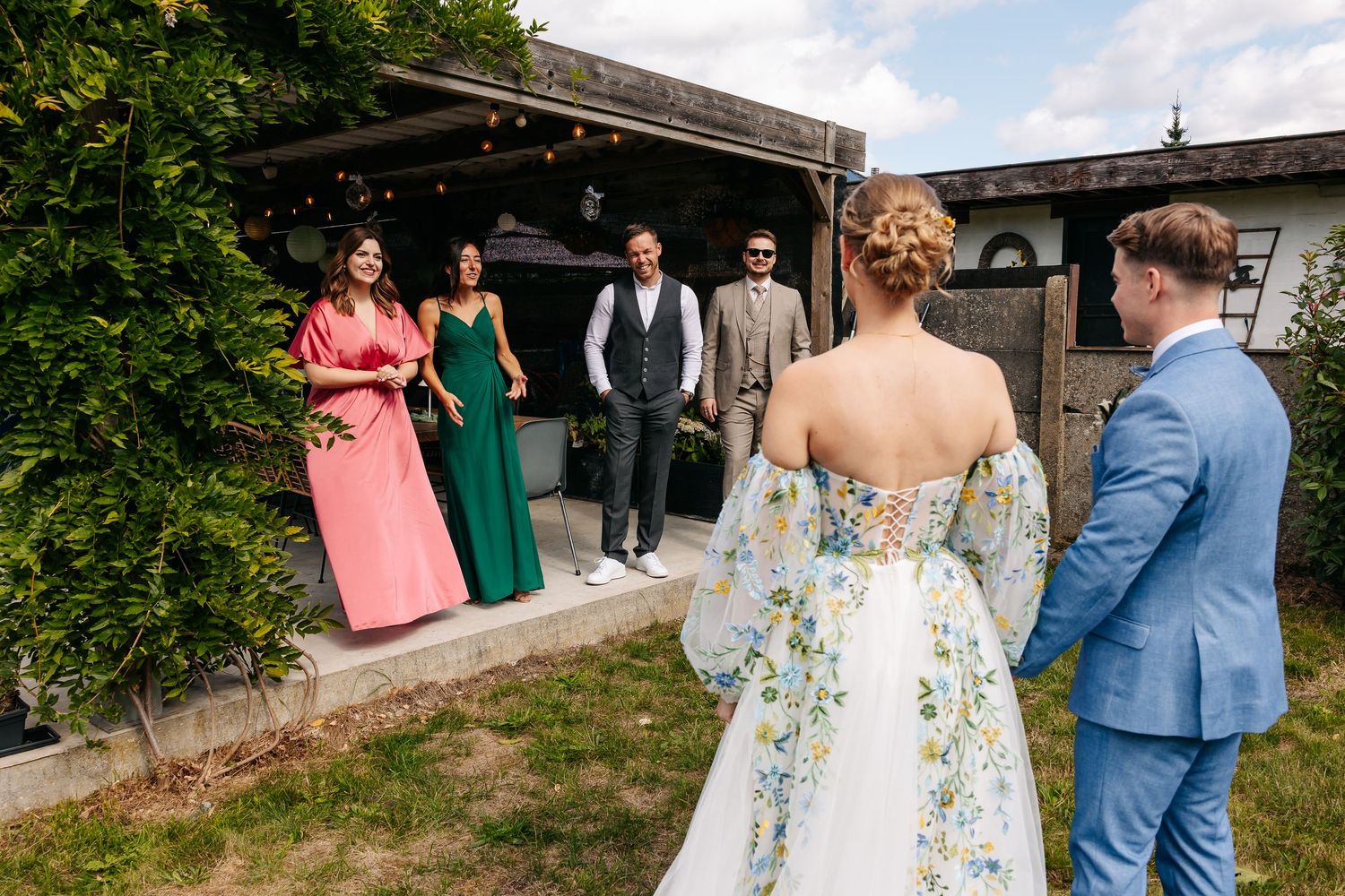 Bride and groom approach smiling wedding party guests gathered outside rustic venue.