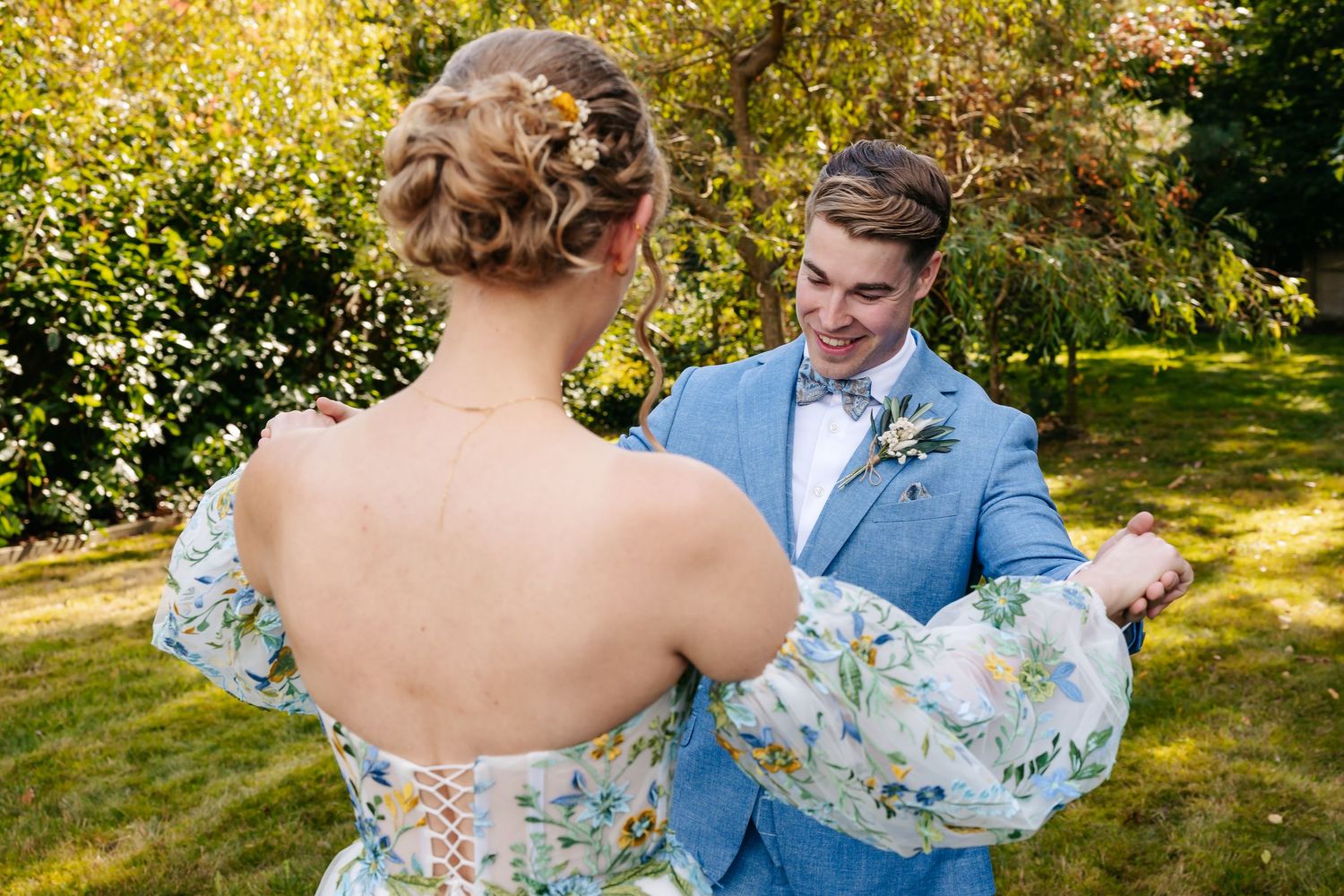 Groom smiles at bride in floral off-shoulder gown with lace-up back during outdoor first look moment.