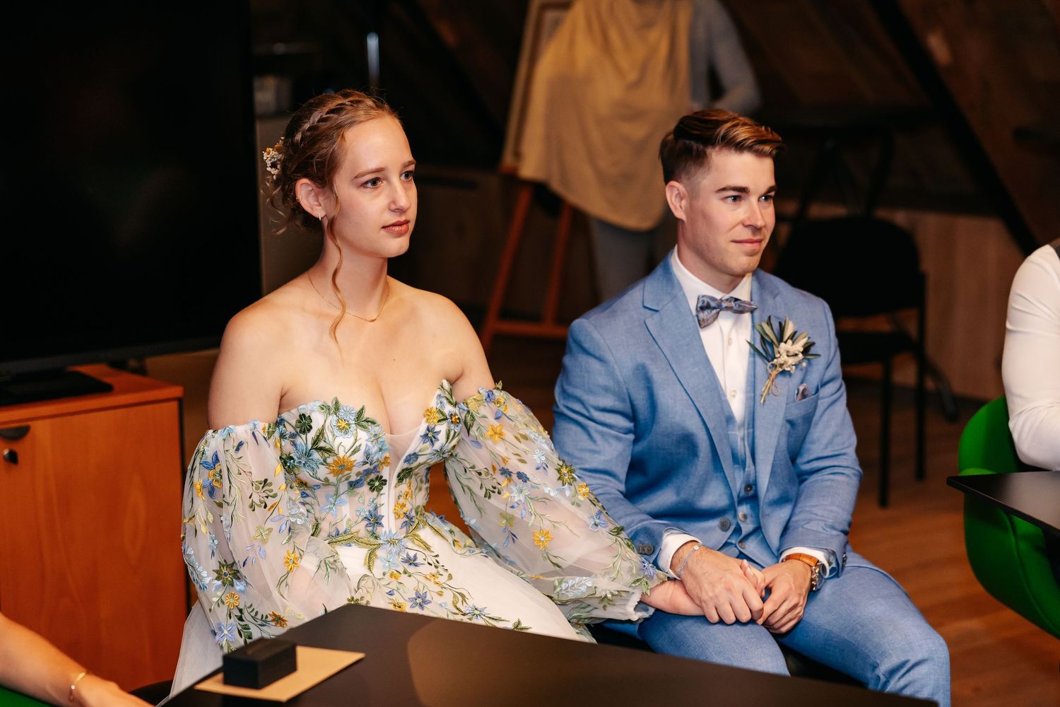 Bride in off-shoulder floral gown sits beside groom in blue suit at ceremony.