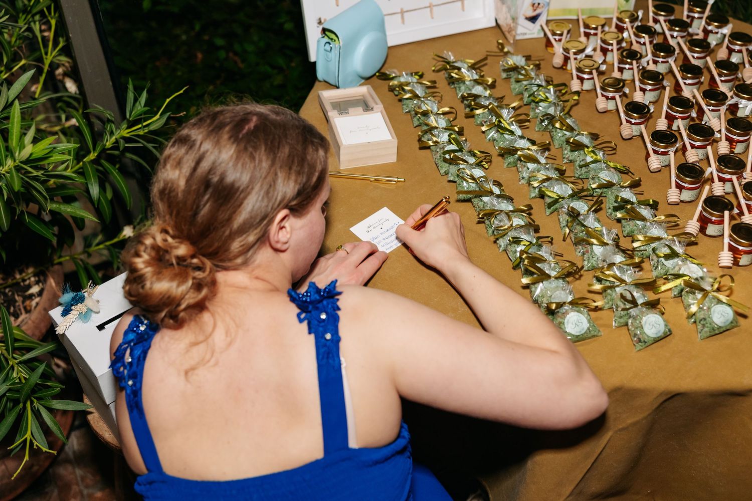 Bridesmaid in blue dress writes place cards at a table displaying floral wedding favors.
