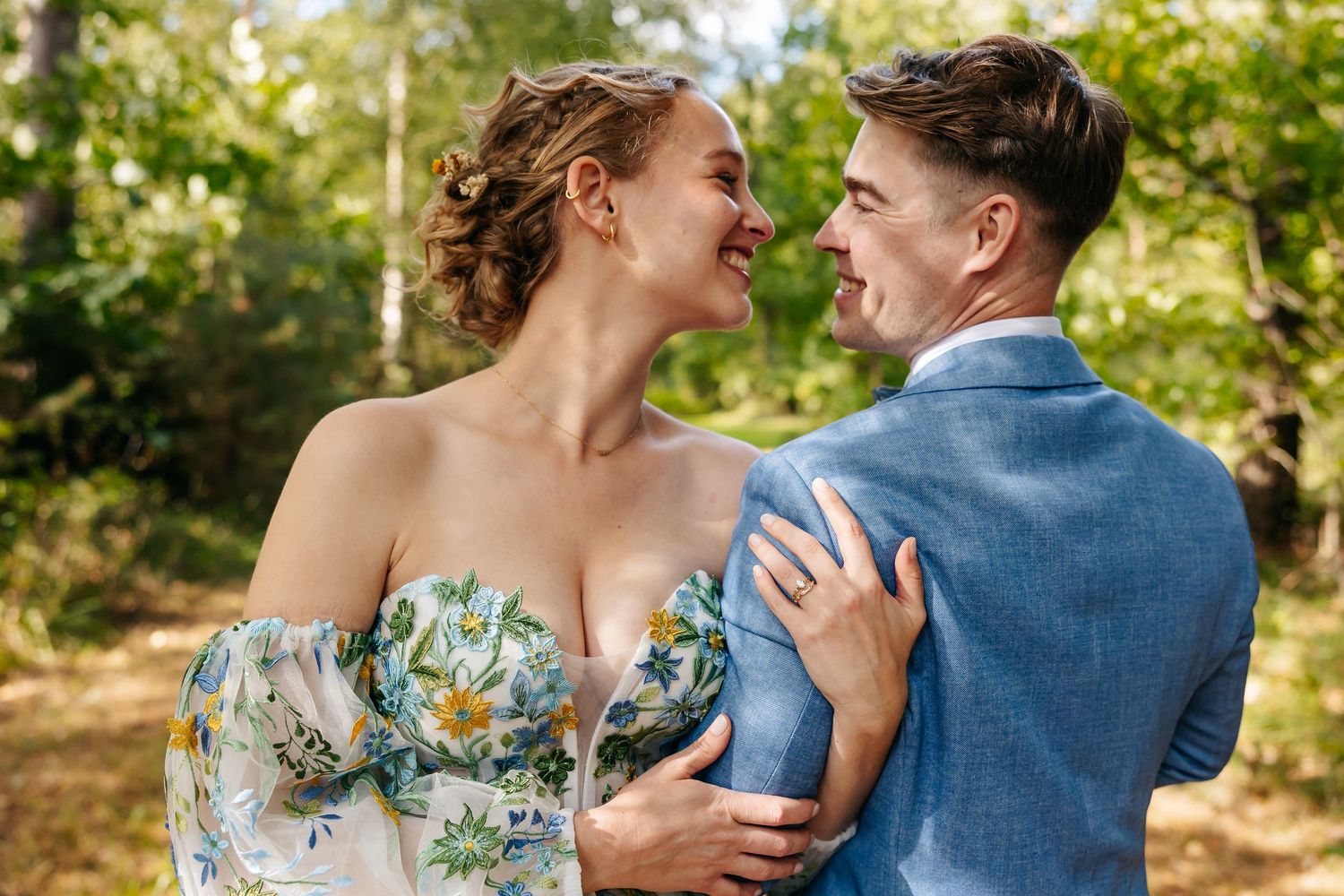 Smiling bride and groom gazing at each other in a lush green forest, bride wearing floral dress and groom in blue suit.
