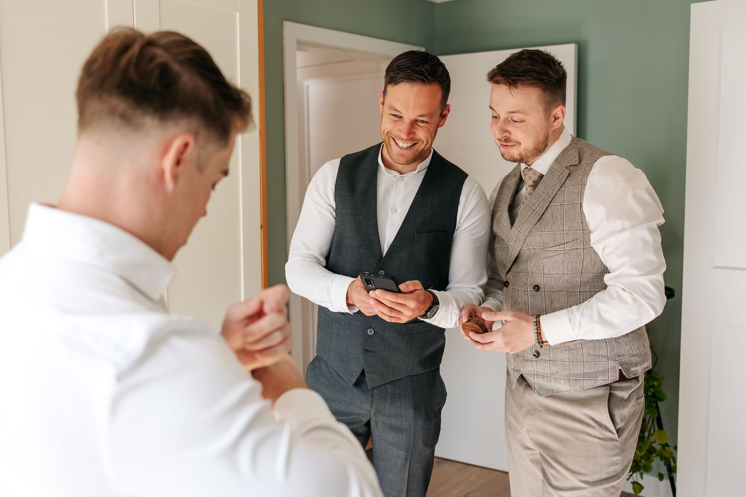 Three groomsmen in suits laughing and looking at wedding rings during pre-ceremony preparations.