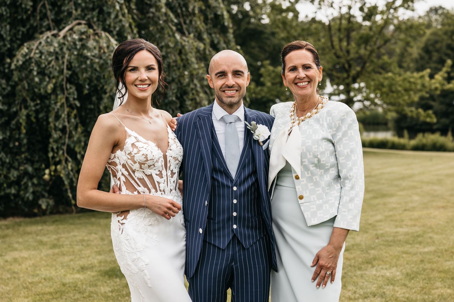 Bride in lace wedding gown and groom in navy suit posing with a woman in a light blue jacket outdoors.