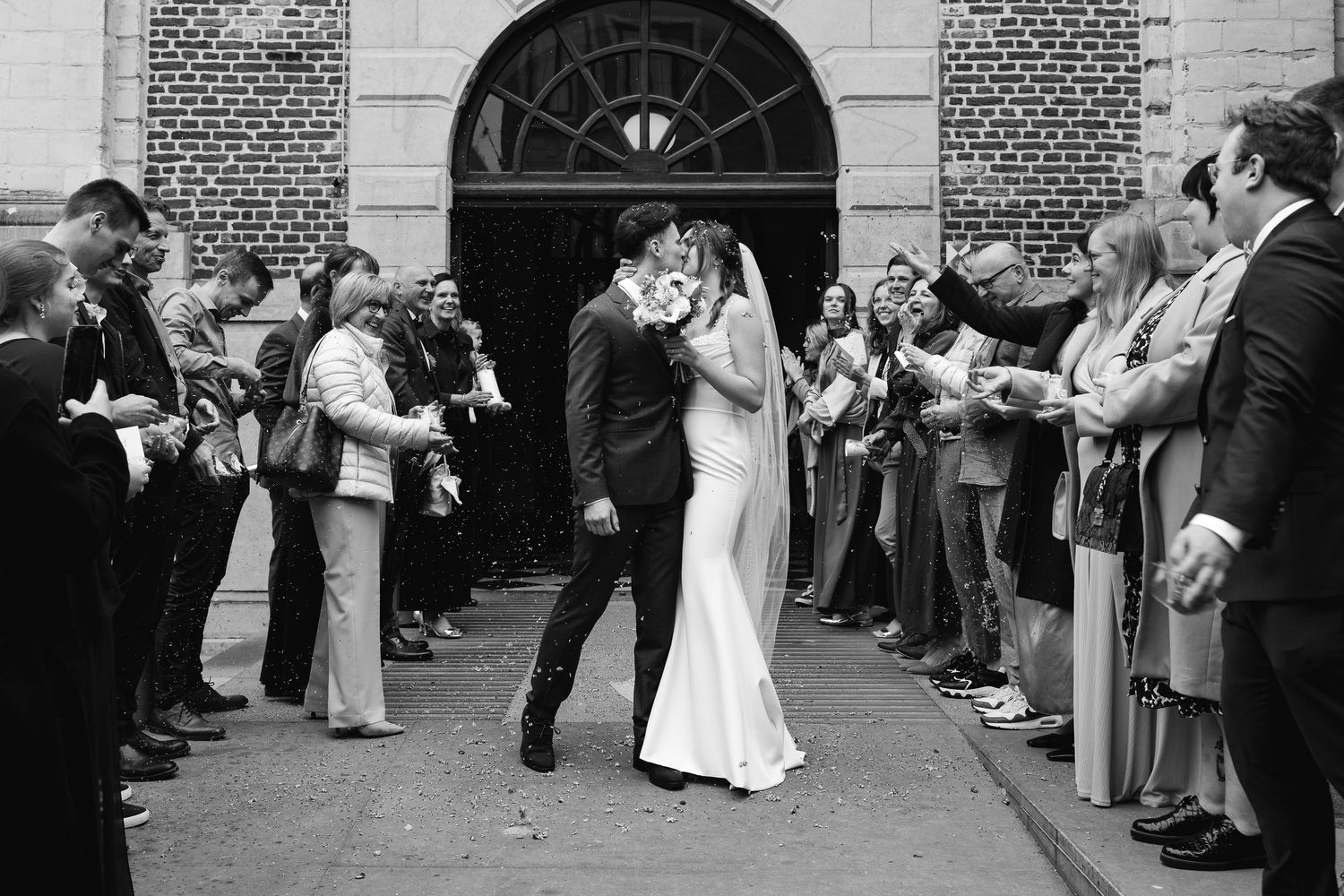 Black and white photo of bride and groom kissing outside church amid celebrating guests.