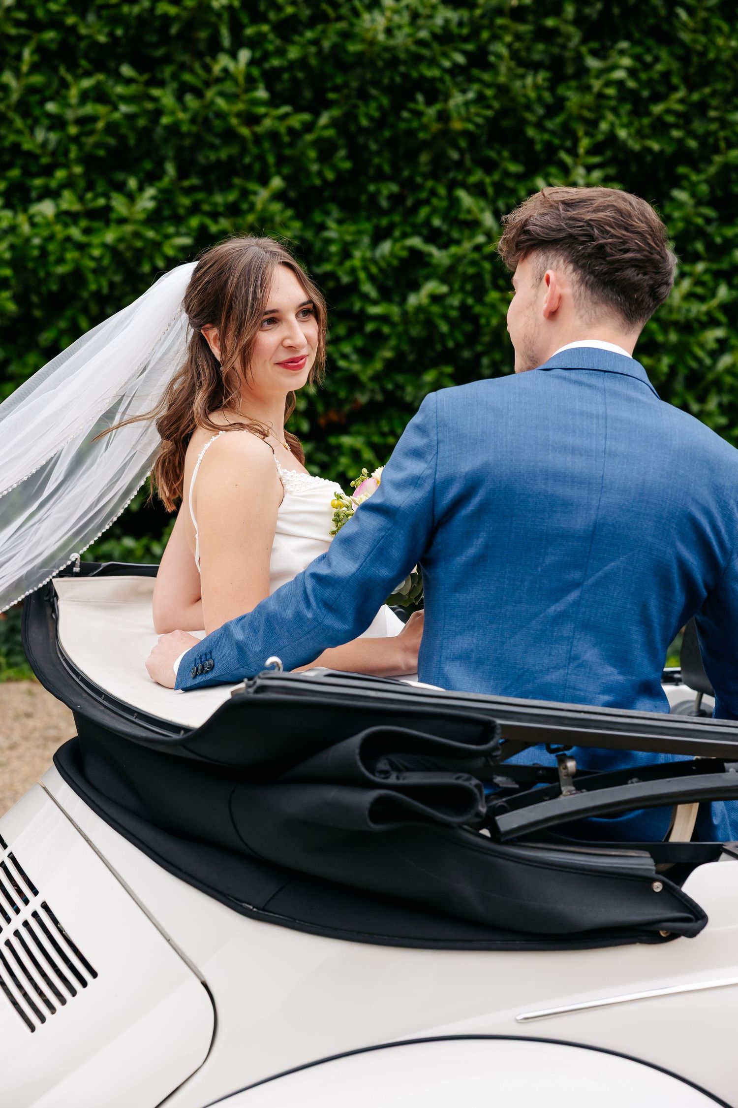 Bride in white veil smiles at groom in blue suit seated in white convertible VW Beetle wedding car.