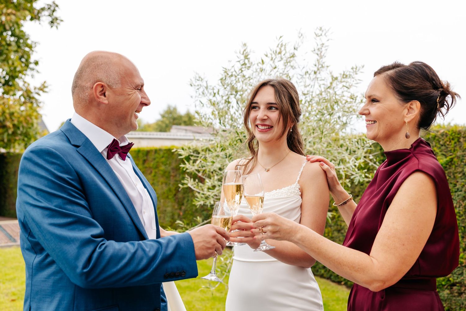 Bride and two guests in burgundy attire toast with champagne glasses at an outdoor wedding celebration.